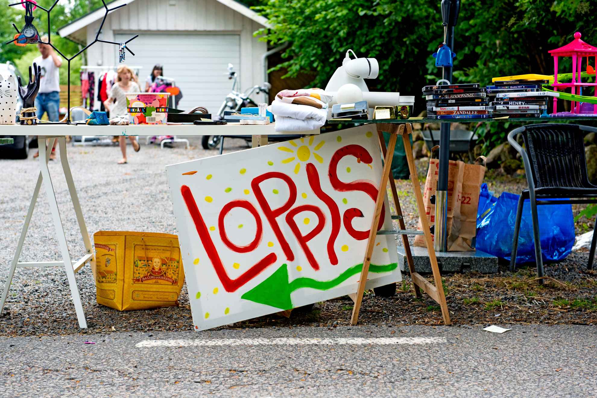 Ein Schild lehnt an einem Tisch. Auf dem Schild steht das Wort Loppis. mit einer Auswahl an gebrauchten Gegenständen. Im Hintergrund ist ein Haus zu sehen.