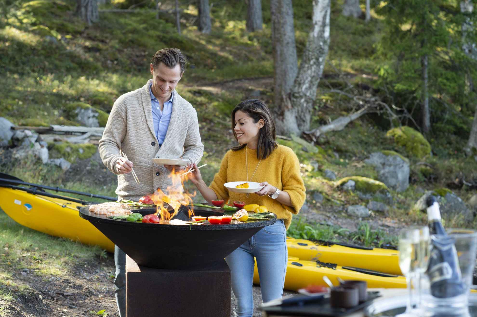 Ein Mann und eine Frau kochen an einem großen Ofen im Freien. Eine Weinflasche im Vordergrund und Kajaks und Wald im Hintergrund.