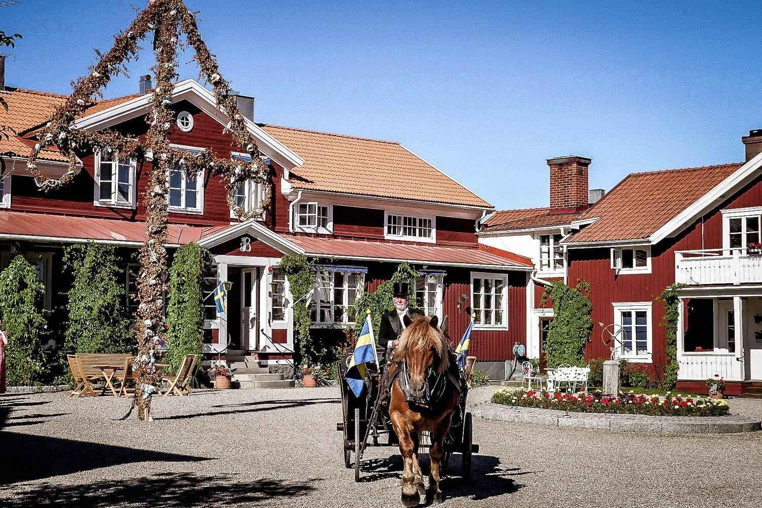 Ein großes rotes Holzhaus mit weißen Rändern im Sommer. Vor dem Haus stehen ein Mittsommerpfahl und ein Pferd mit Wagen mit zwei schwedischen Fahnen.