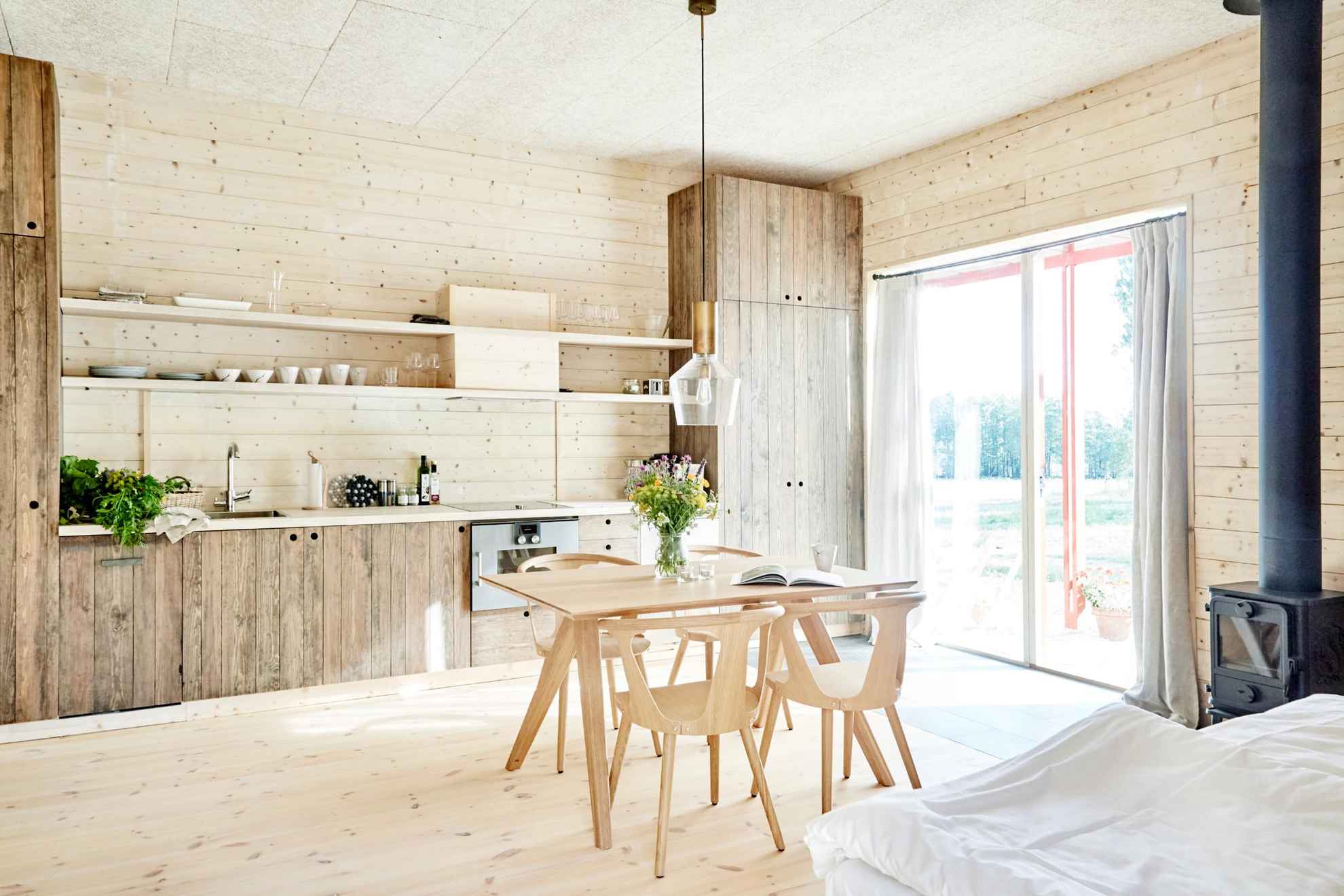 Ein Zimmer mit einer Küche, Bett, Kamin, Küchentisch und Stühlen. Der Raum hat einen Holzboden und Wände, und zwei Glastüren führen auf eine Terrasse.