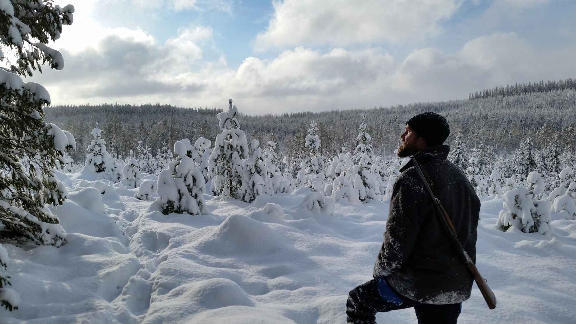 In einem verschneiten Wald steht ein Mann mit einem Jagdgewehr auf dem Rücken.