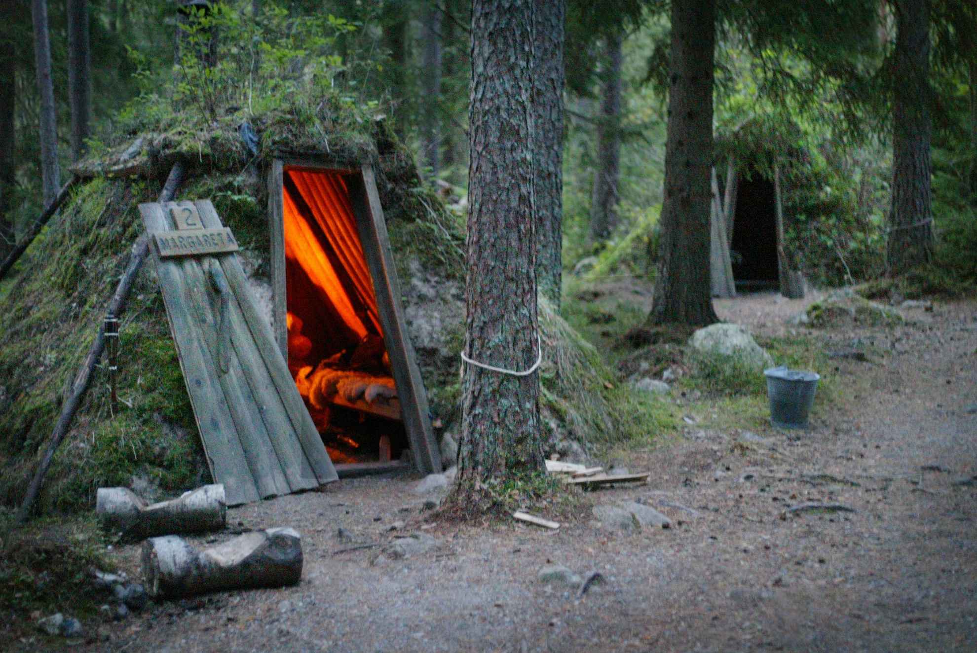 Zwei mit Moos bedeckte Waldhütten in einem Wald. In einer der Hütten brennt ein Kamin.