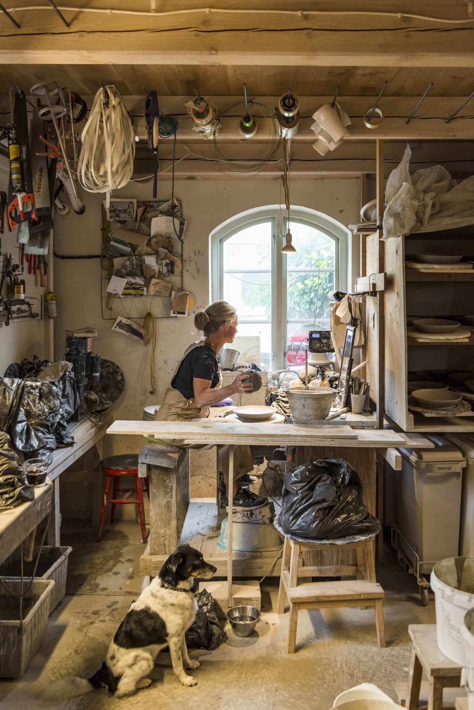Eine Frau sitzt an einem Fenster und arbeitet in ihrem Töpferatelier. Es gibt viele Werkzeuge und Keramik im Raum. Neben ihr sitzt ein Hund auf dem Boden.