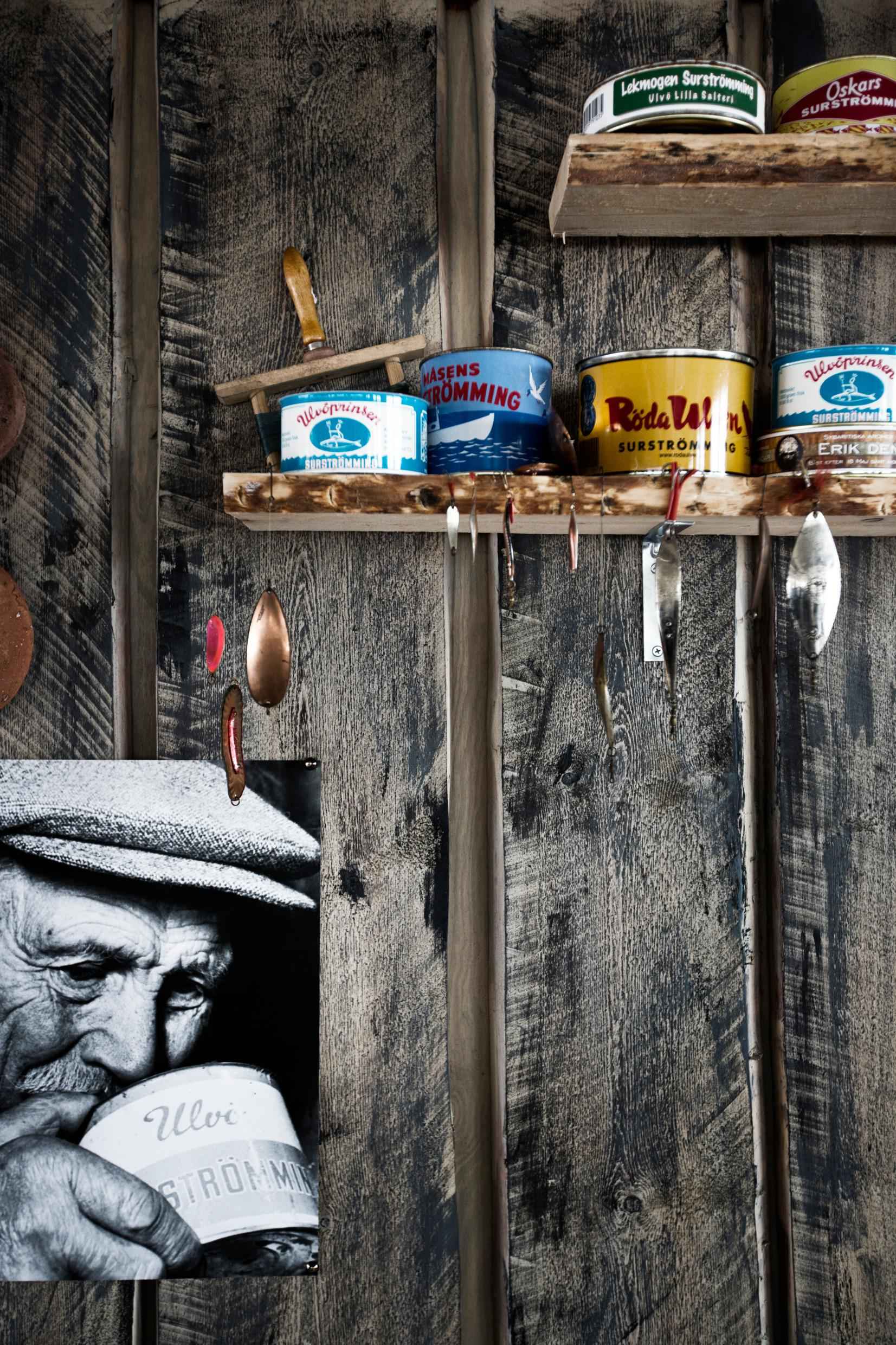 An einer Holzwand stehen verschieden Dosen mit fermentiertem Hering auf einem Regal. Ein schwarz-weißes Foto eines alten Mannes hängt an der Wand., der an einer Dose riecht.