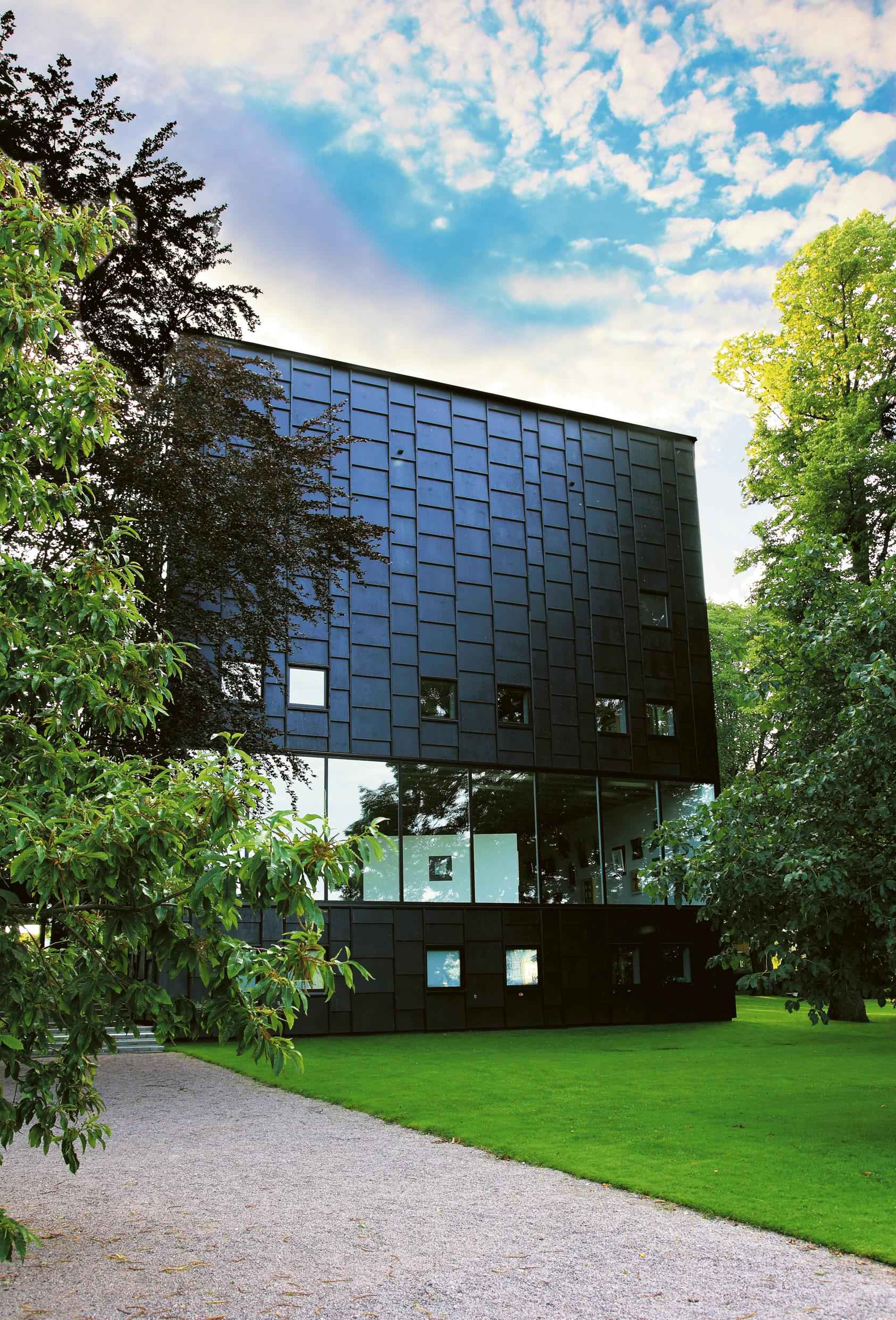 Äußeres des Kunstmuseums Kalmar, eines modernes schwarzen Gebäudes mit einer Reihe großer Fenster, in denen du die Kunstausstellung sehen kannst. Zwei große Bäume und eine grüne Wiese vor dem Gebäude.