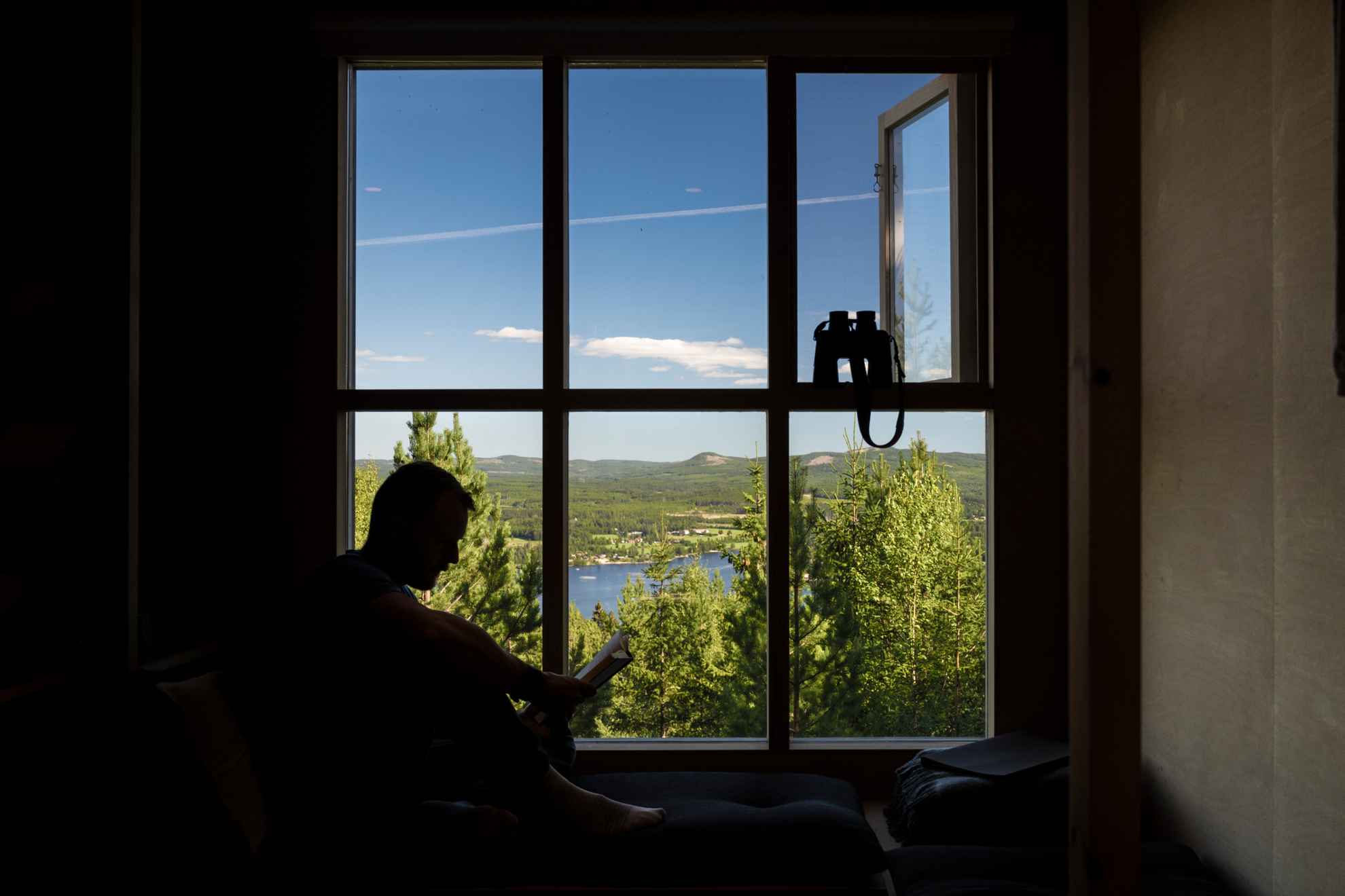 Silhouette eines lesenden Mannes vor einem großen Fenster mit Blick auf den blauen Himmel und die Berge in Hälsingland, mit einem Fernglas in der Hand.