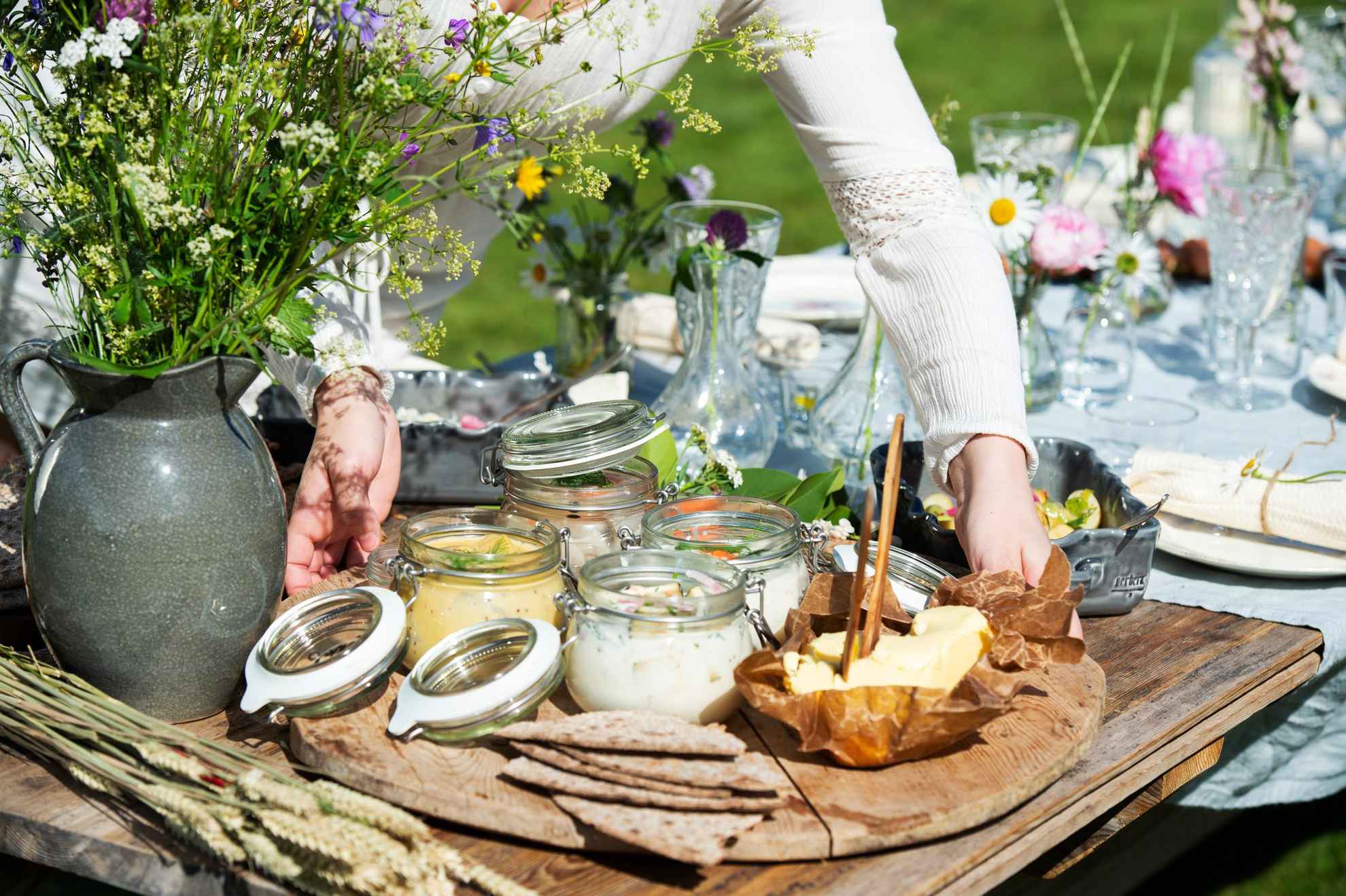 Eine Frau stellt ein Tablett mit eingelegtem Hering, Butter und Brot auf einen für das Mittsommeressen gedeckten Tisch. Neben dem Tablett steht eine Vase mit Wildblumen.