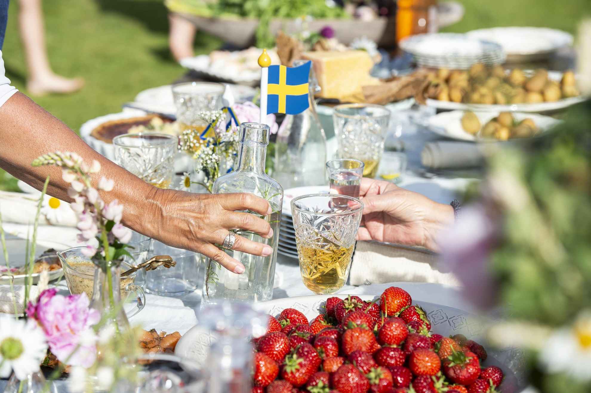 Ein gedeckter Tisch für ein Mittsommer-Mittagessen mit traditionellen Speisen. Eine Hand hält eine Flasche Schnaps und eine andere Hand hält ein Glas Schnaps. Im Vordergrund steht eine Schüssel Erdbeeren.