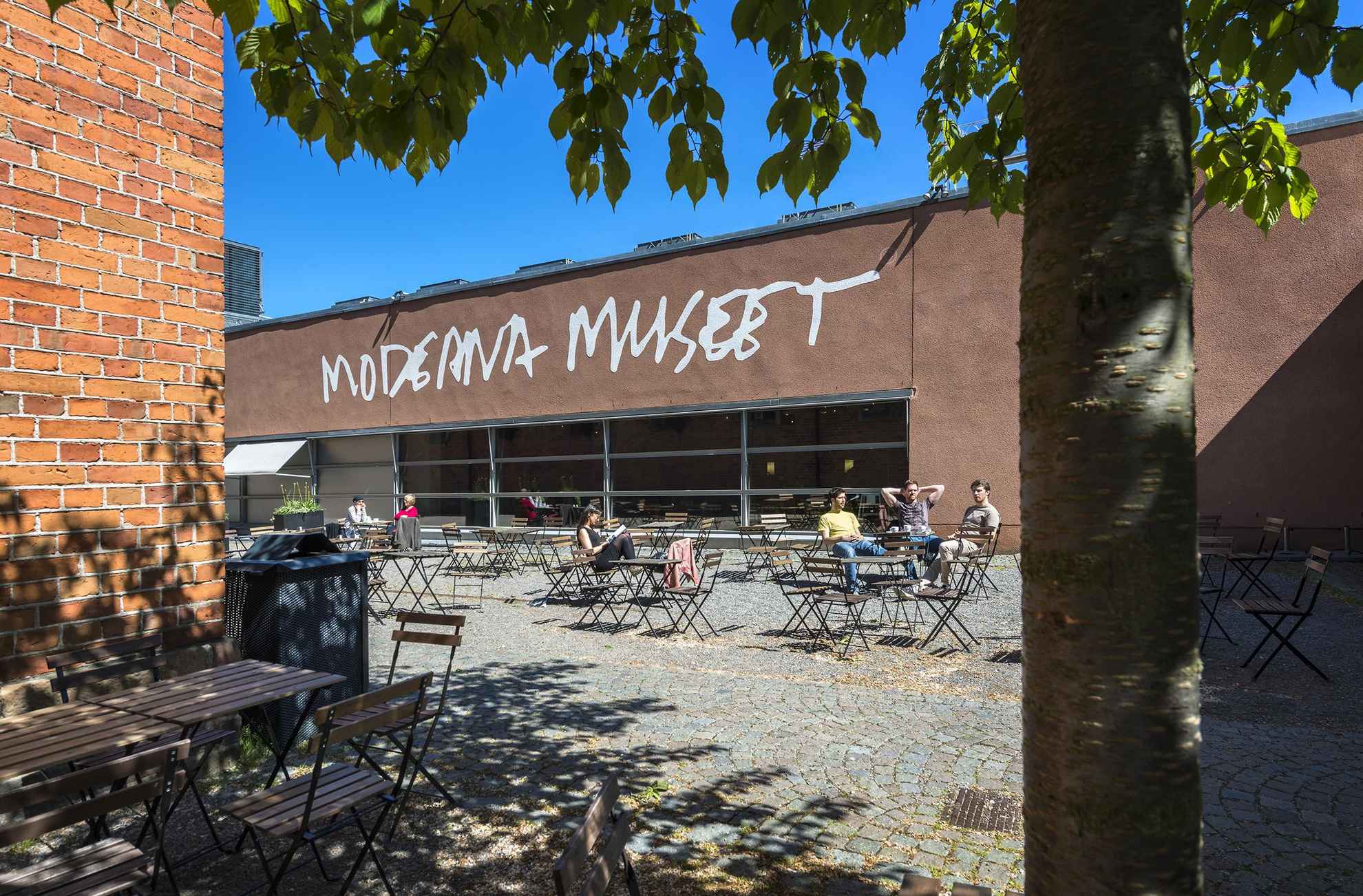 Außenansicht des Moderna Museet in Stockholm.  Vor dem Museum stehen viele Gartenmöbel und Menschen sitzen in der Sonne.