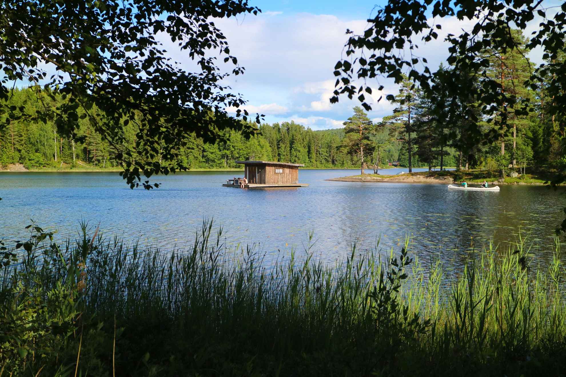 Eine Hütte, die in einem See bei Naturbyn schwimmt, und jemand, der in einem Kanu daran vorbeifährt. Der See ist von Wald umgeben.