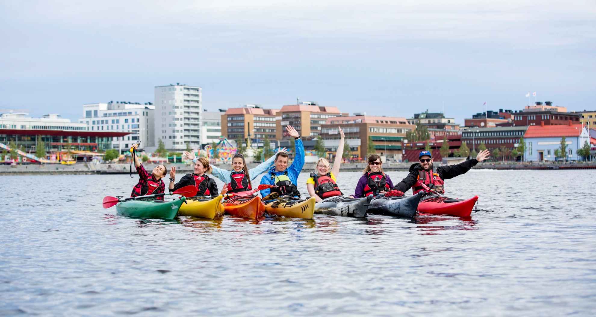 Eine Gruppe von sieben Personen fährt Kajak. Sie sind auf dem Meer vor der Stadt Luleå versammelt und schauen in die Kamera.
