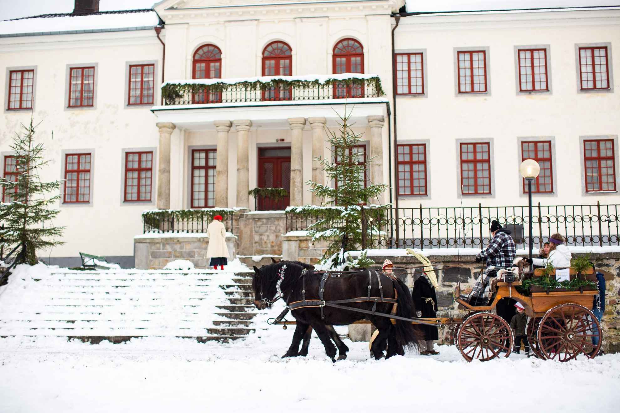 Ein Pferd und eine Kutsche stehen im Winter vor einem großen weißen Haus. Auf dem Boden liegt Schnee, ein paar Menschen stehen neben der Kutsche und ein paar sitzen auf ihr
