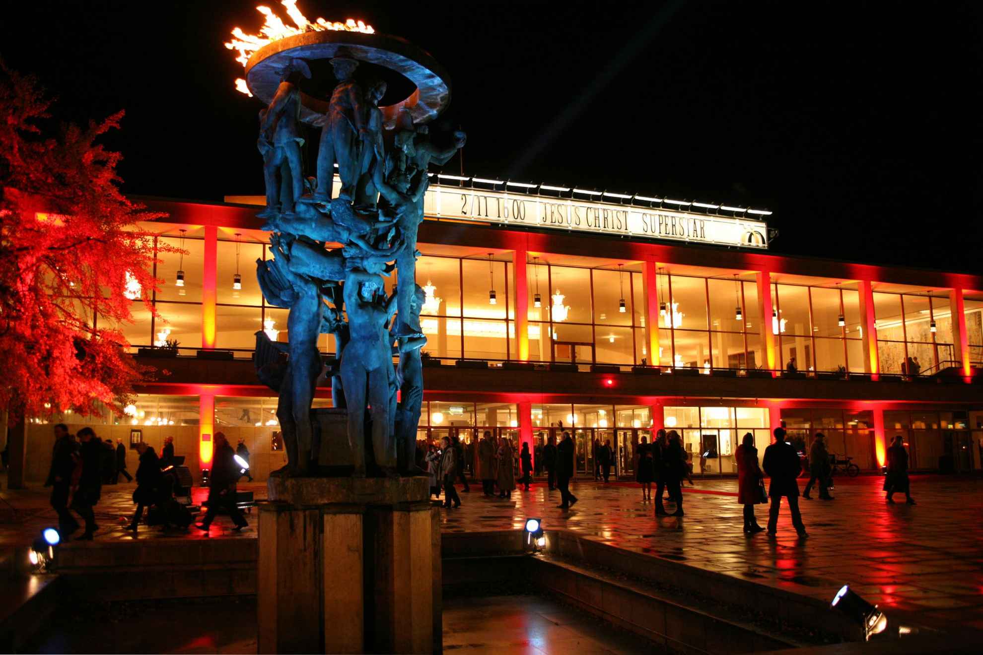 Die Statue, bestehend aus Menschen die einen Feuerring halten steht vor der Malmö Oper, ein großes Gebäude mit Panoramascheiben.