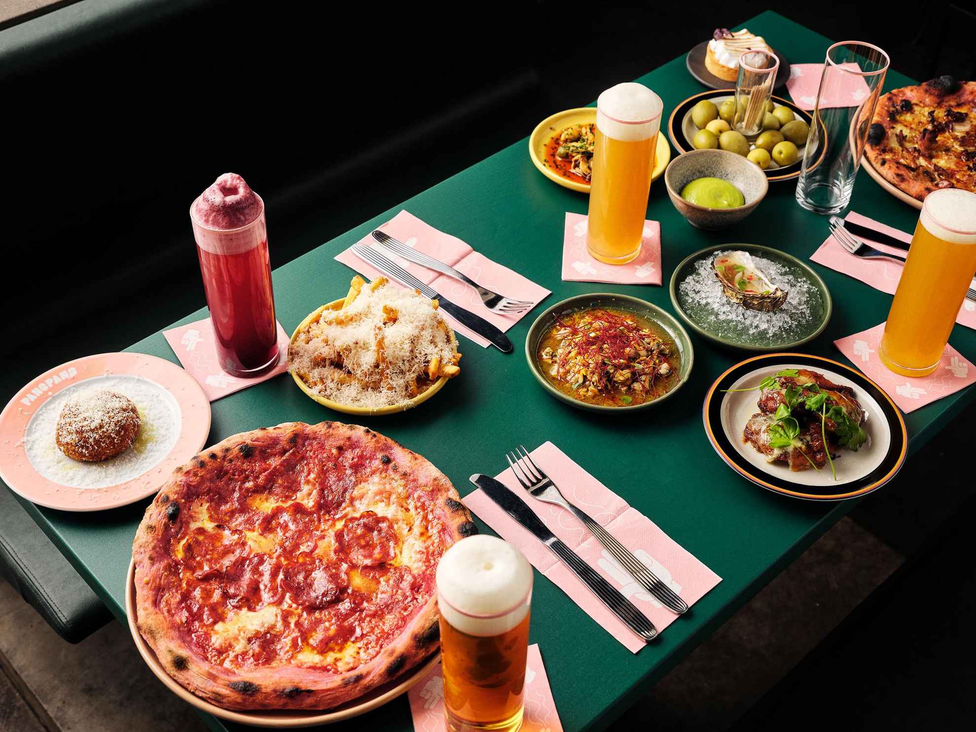 Ein grüner Tisch mit Besteck, mit Bier gefüllten Gläsern und Tellern mit Speisen wie Pizza, Pommes frites, Austern und Oliven.
