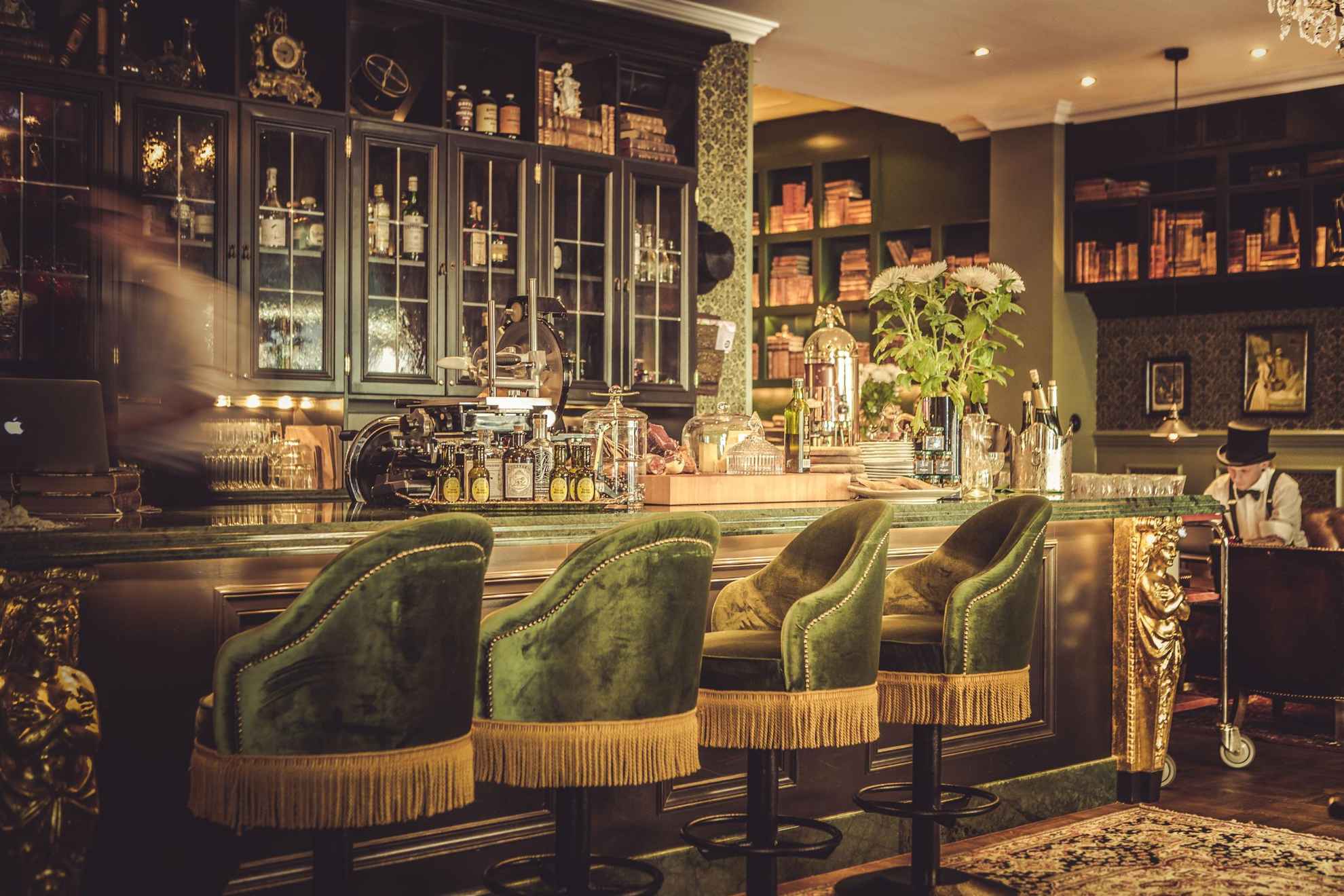 Die Bar im Hotel Pigalle, mit grünen Samtstühlen, Holzregalen und goldenen Details. Jemand bewegt sich hinter der Bar.
