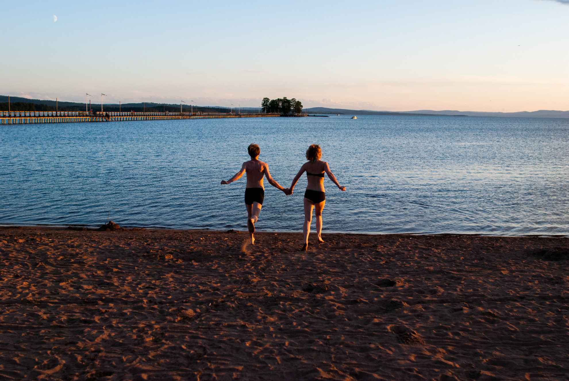 Zwei Personen in Badeanzügen laufen an einem Strand in Richtung Wasser.