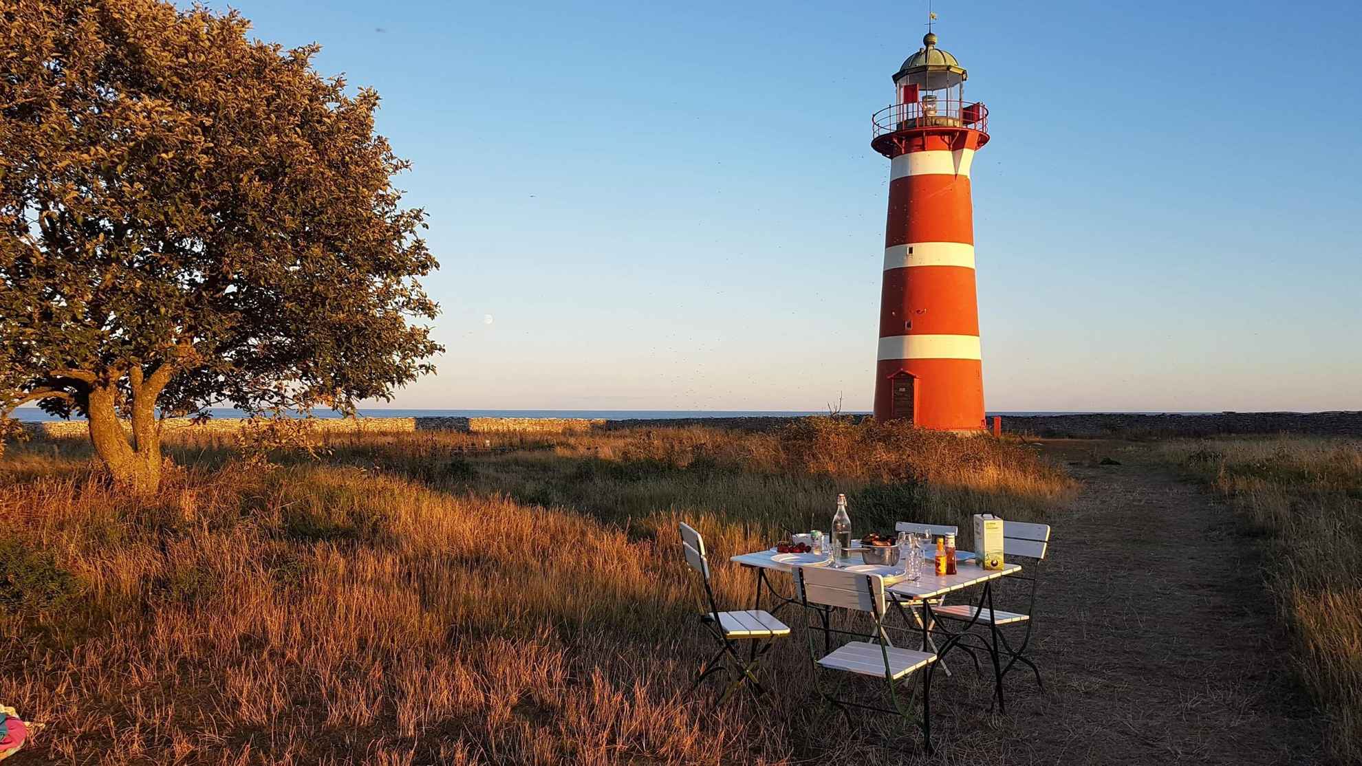Ein Holztisch mit vier Stühlen steht im Sommer im Freien. Im Hintergrund ist ein rot-weiß gestreifter Leuchtturm zu sehen.