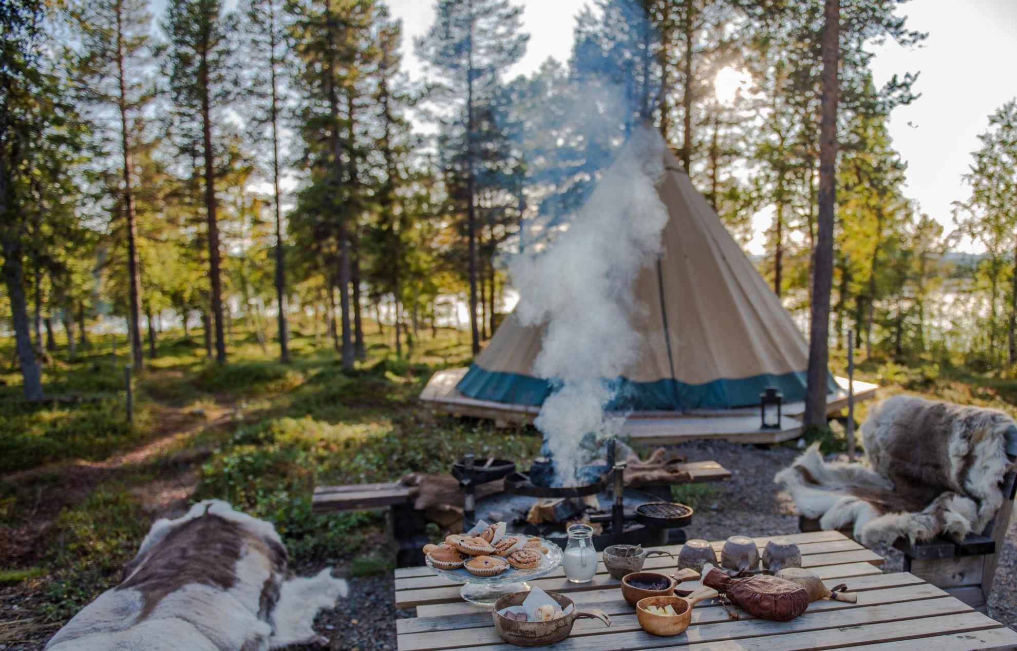 Mit Rentierfellen bezogene Bänke neben einem Tisch mit Tassen und Schüsseln und einem Teller mit Brötchen. Eine Kaffeekanne wird über ein Lagerfeuer gestellt. Ein Lavvu-Zelt im Hintergrund und man sieht einen See zwischen den Bäumen.