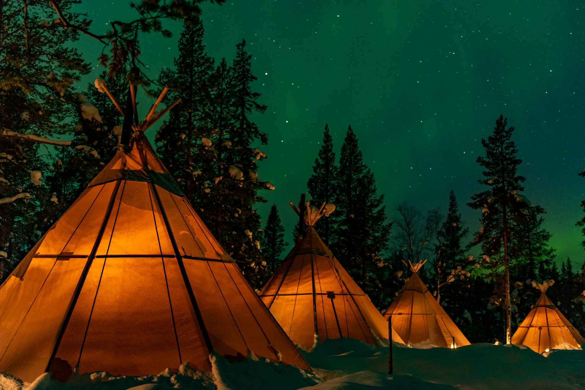 Vier Lavvu-Zelte in einem Naturcamp in Lappland. Es ist eine sternenklare Nacht in einer verschneiten Landschaft und das Licht glüht in den Zelten.