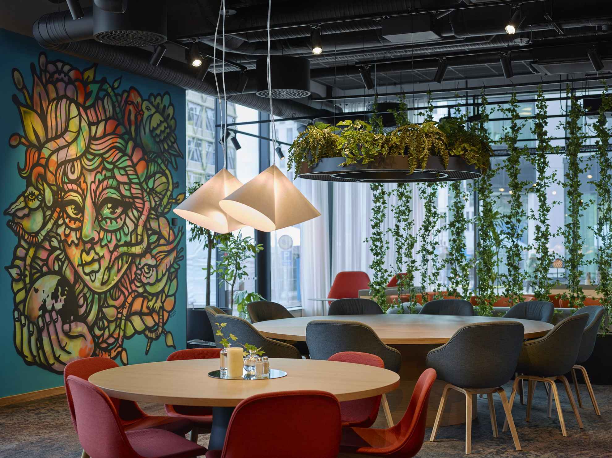 Ein Raum mit runden Holztischen und roten und schwarzen Stühlen. Pflanzen hängen von der Decke und eine blaue Wand ist mit Kunstwerken bemalt.