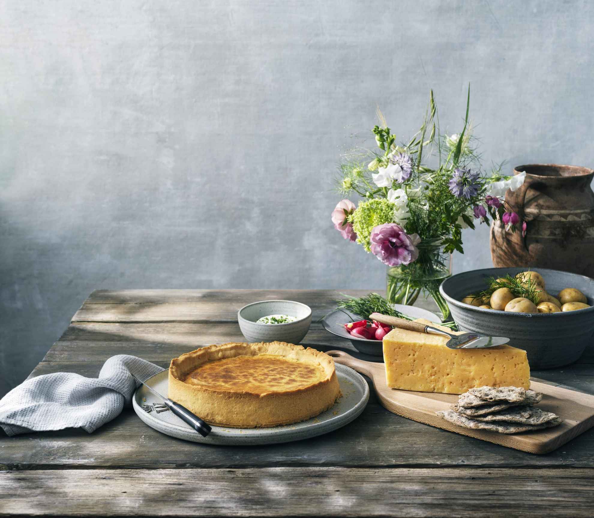Ein Holztisch mit Västerbottenpaj-Quiche, Västerbotten-Käse, einer Schüssel Kartoffeln und einer Blumenvase.