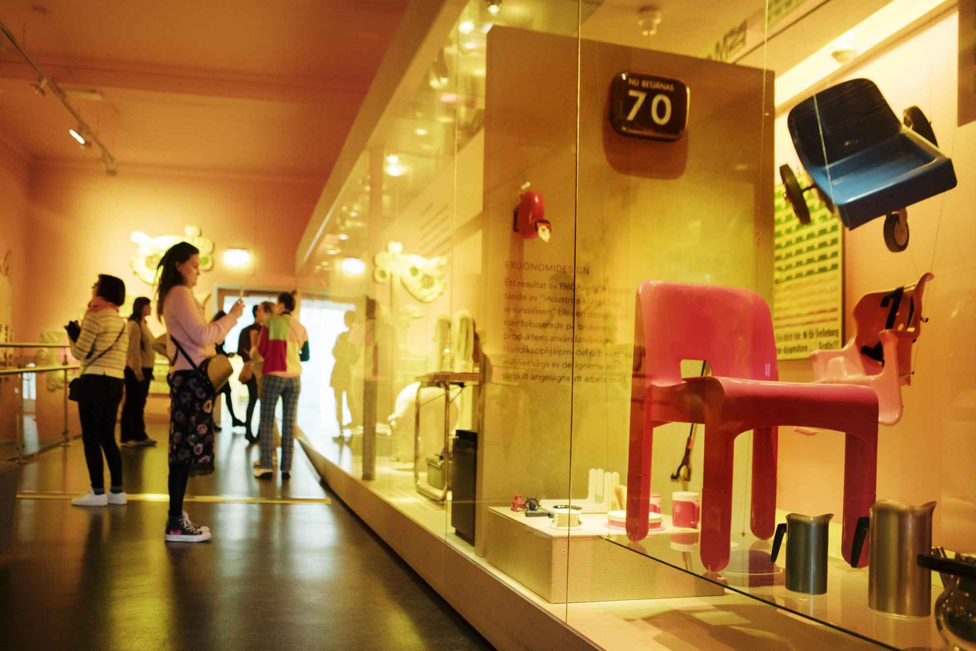 Besucher des Design und Handwerks Museums betrachten die Ausstellung mit Design Artikeln wie einen roten Stuhl.