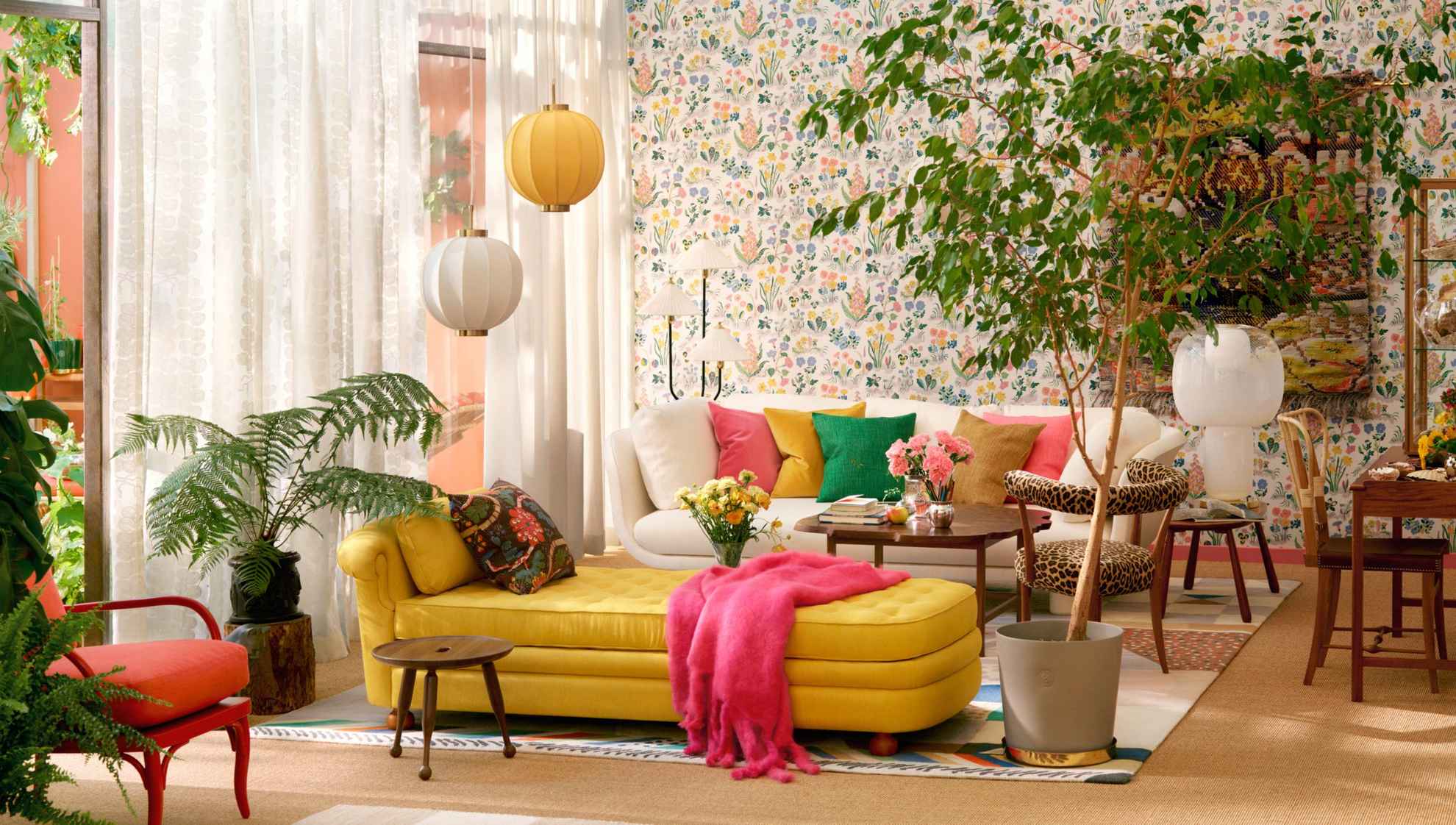 Ein Wohnzimmer mit einem beigen Sofa mit bunten Kissen im hinteren Bereich und einer gelben Chaiselongue in der Mitte des Raums. Rechts von der Chaiselongue steht eine große grüne Bodenpflanze. Die durchsichtigen weißen Vorhänge lassen natürliches Licht herein.