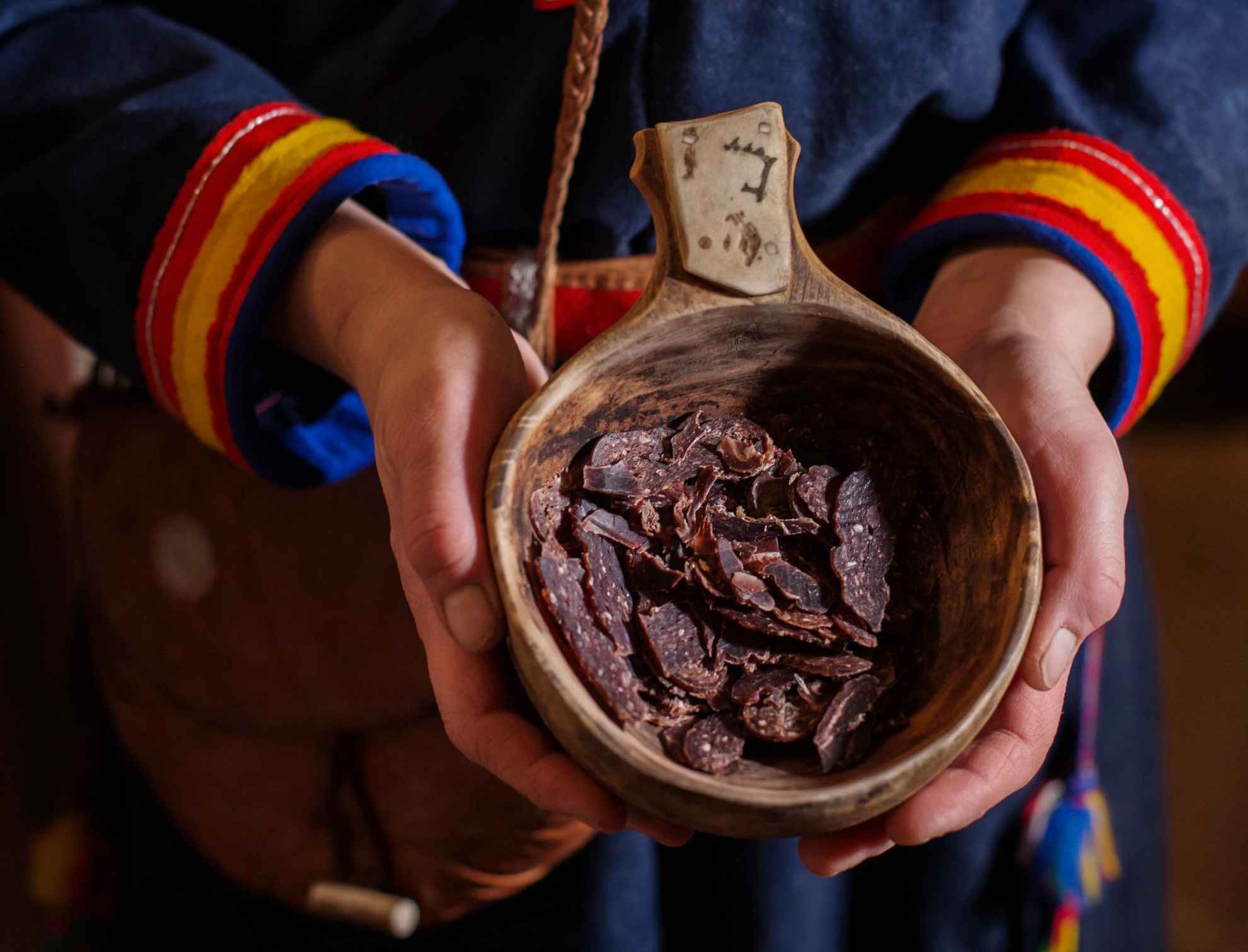 Eine Holzschale mit Rentierfleisch in der Hand einer Person die samische Kleidung trägt.