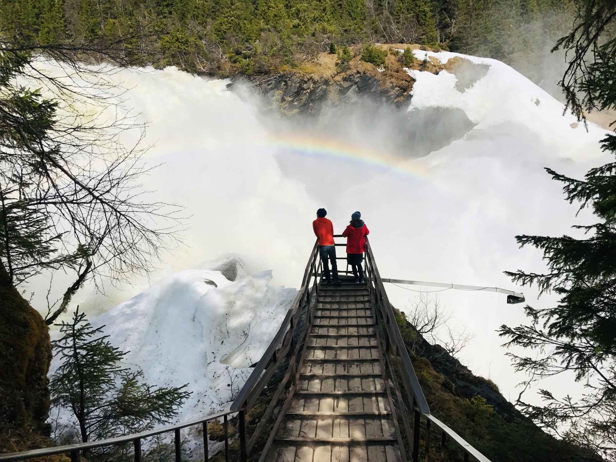 Zwei Personen blicken auf einen riesigen Wasserfall. Das Wasser des Wasserfalls erzeugt einen Regenbogen.