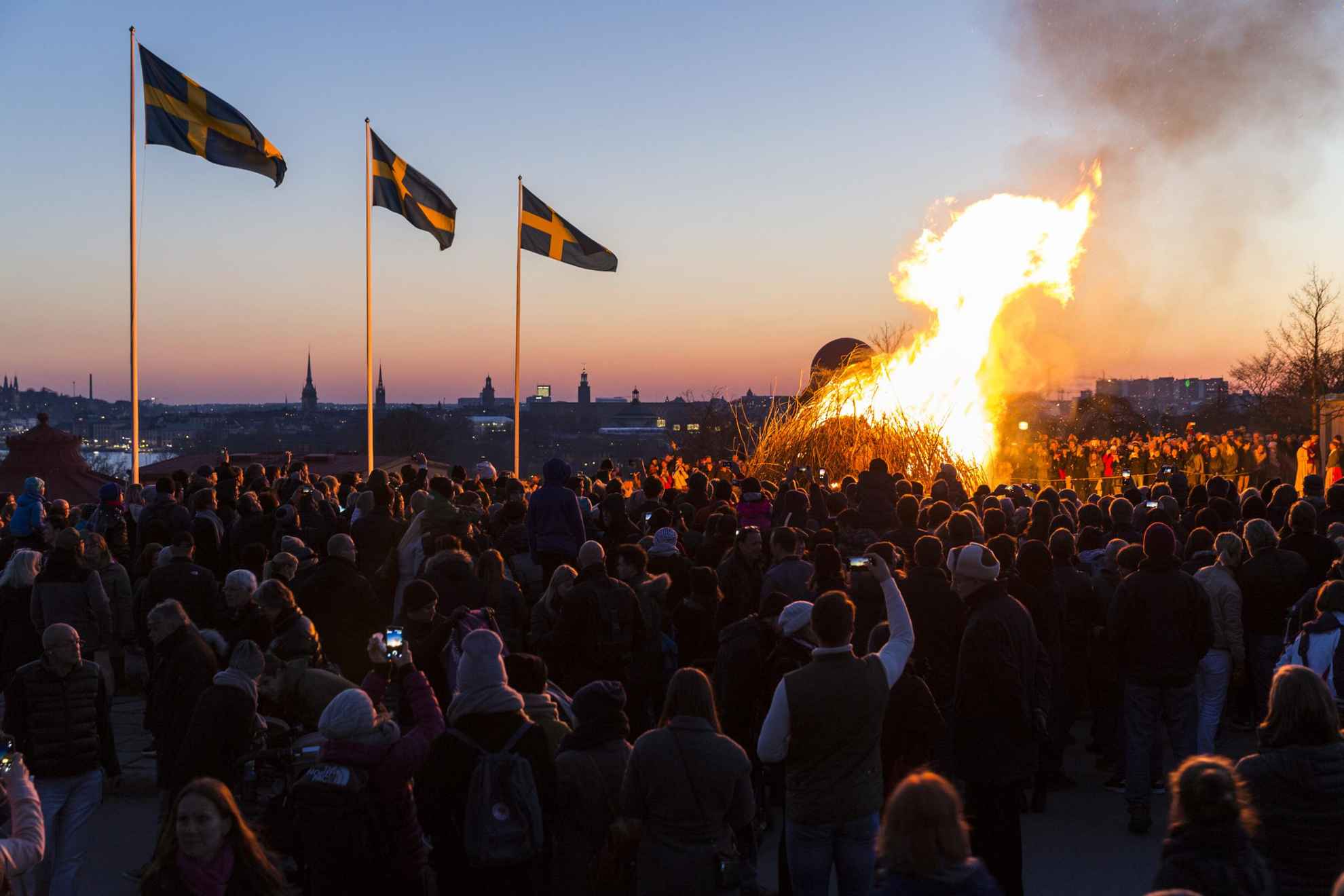 Menschen haben sich um ein Lagerfeuer versammelt, um die Walpurgisnacht zu feiern. Drei schwedische Fahnen wehen im Wind.