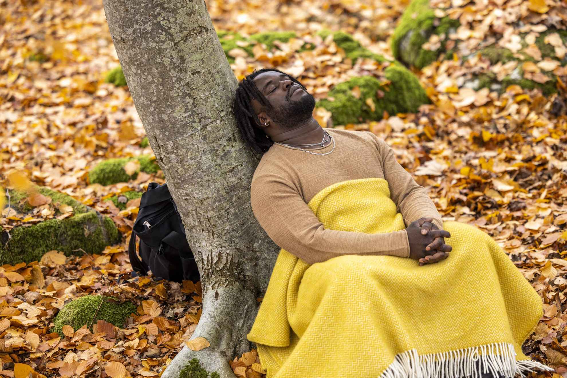 Ein Mann lehnt im Herbst mit geschlossenen Augen an einem Baum. Er hat eine gelbe Decke über seinen Beinen und um ihn herum liegen Blätter auf dem Boden.