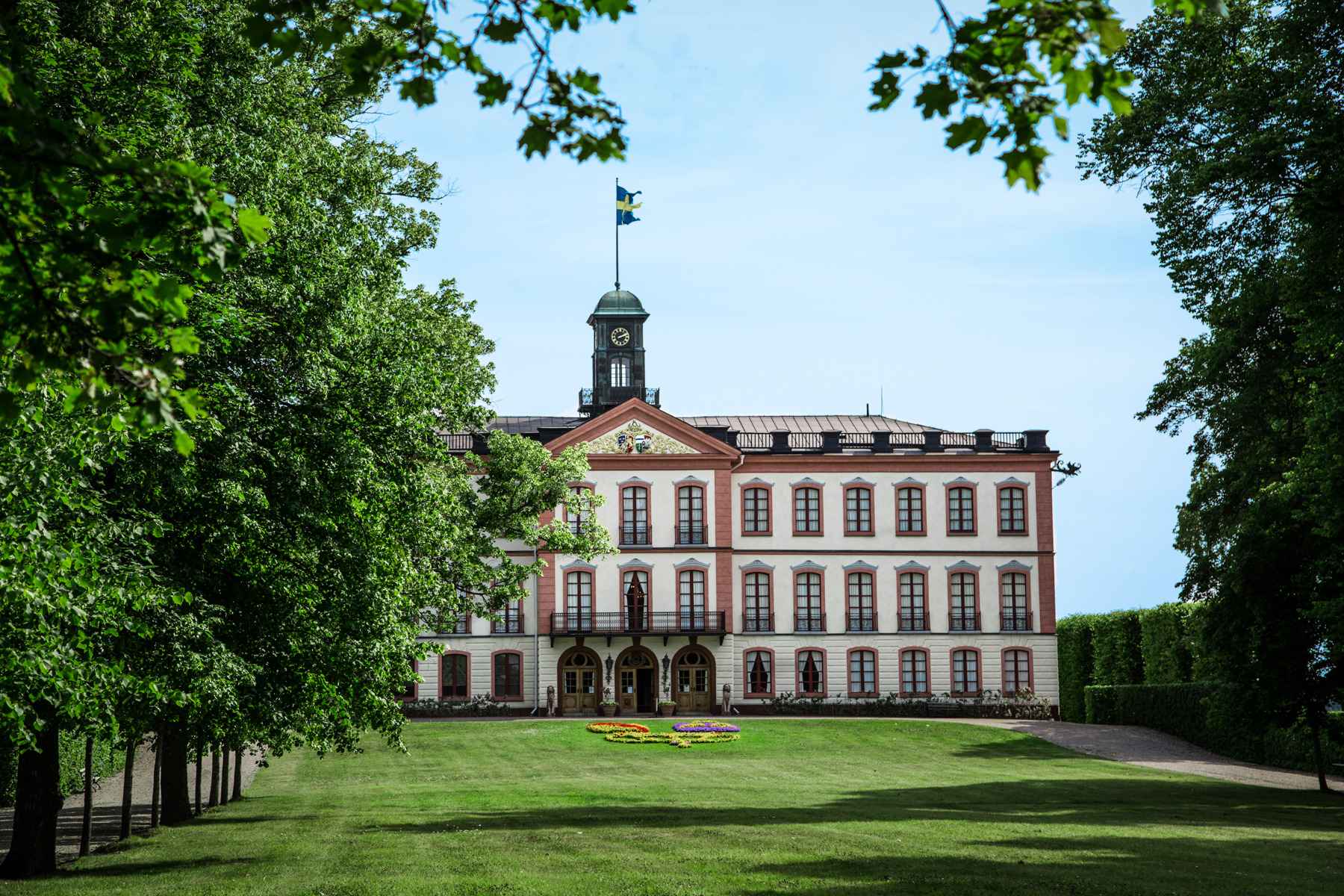 The exterior of Tullgarn Castle which has white walls and large windows surrounded with red details. Trees and a lawn surrounds the building.