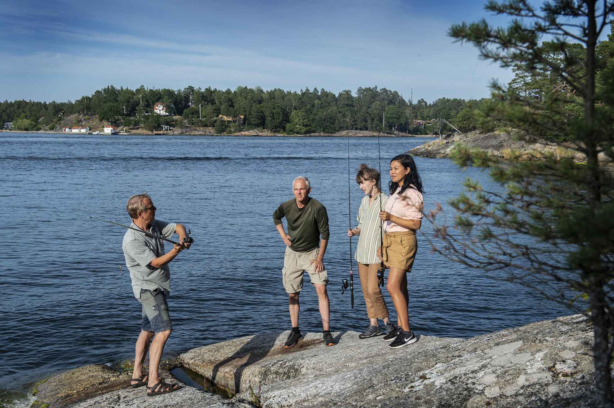 A man is showing another man and two women how to fish on a cliff in the archipelago.
