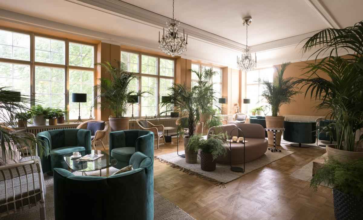 Eine Sitzecke mit Sofas und Sesseln. Der Raum ist mit Pflanzen und zwei von der Decke hängenden Kronleuchtern dekoriert.