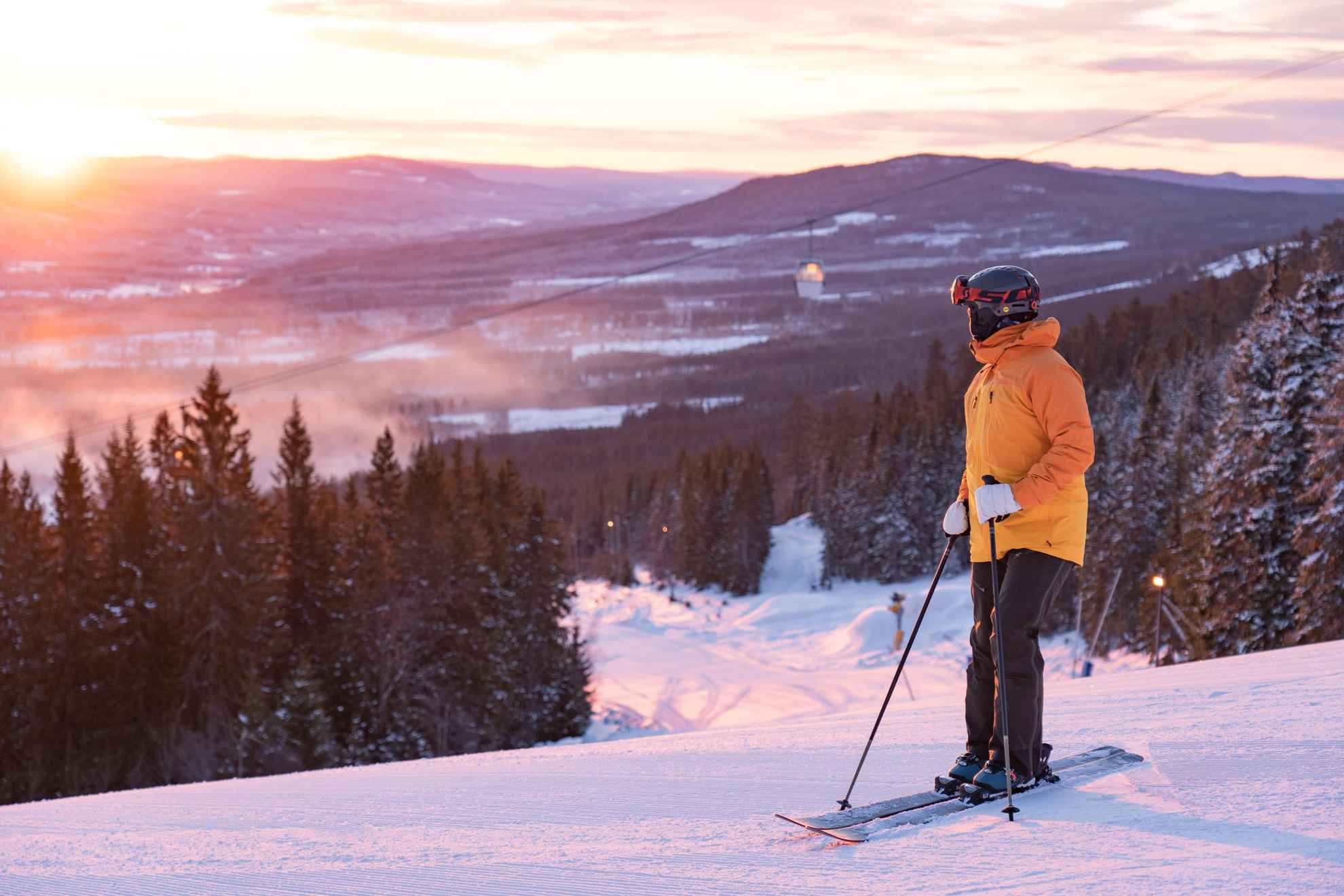 Eine Person mit Skiausrüstung steht auf einer Skipiste und betrachtet den Sonnenuntergang.