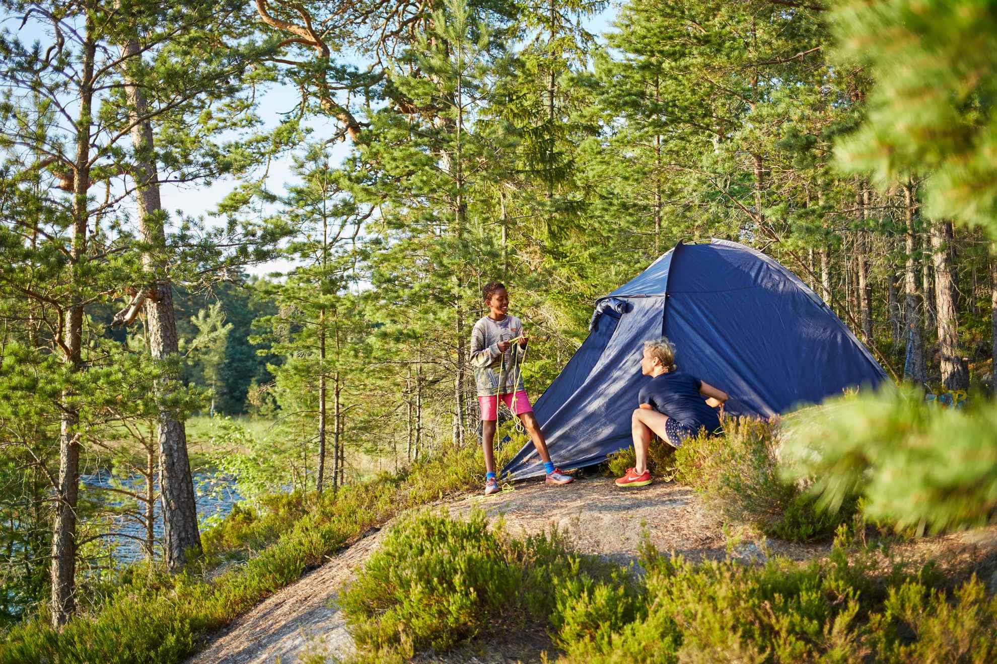 Zwei Menschen in Schweden stehen bei einem Zelt im Wald an einem See.