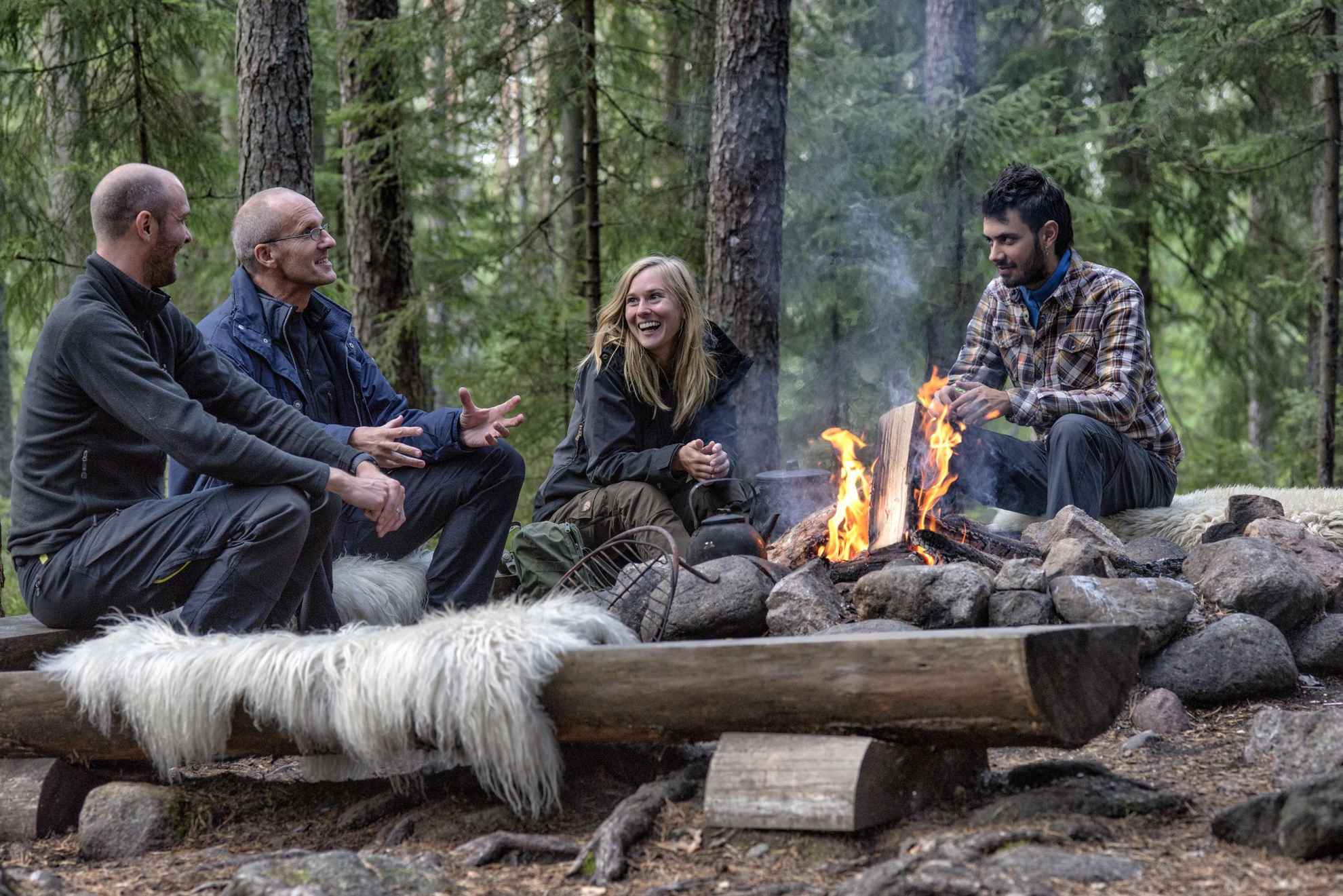 Vier Leute sitzen lachend um ein Lagerfeuer in einem Wald.