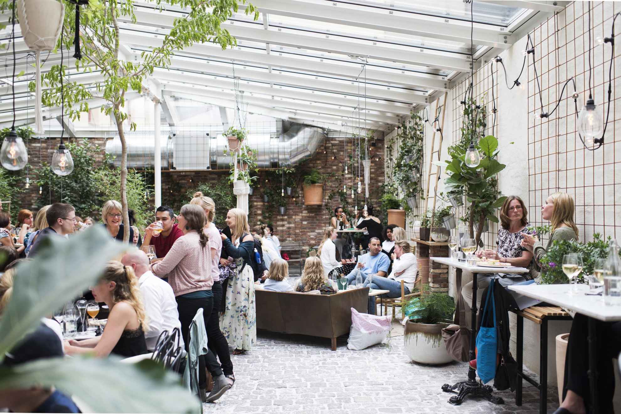 Menschen genießen Getränke und Speisen in dem belebten Café Magasinet in Göteborg. Der Innenraum besteht aus Steinboden, Backsteinwänden, einem Glasdach und der Raum ist voller grüner Pflanzen.