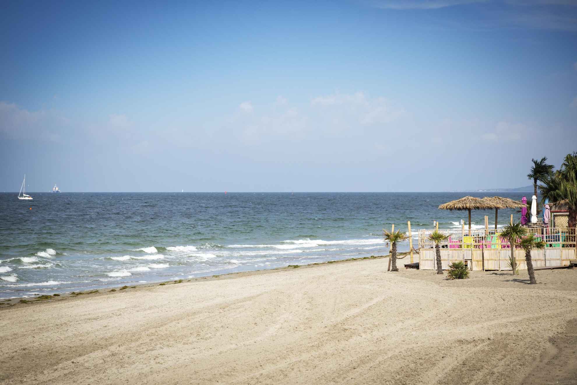 Ein aufgeräumter Sandstrand am Meer. Rechts stehen Palmen und Sonnenschirme mit Strohdächern.