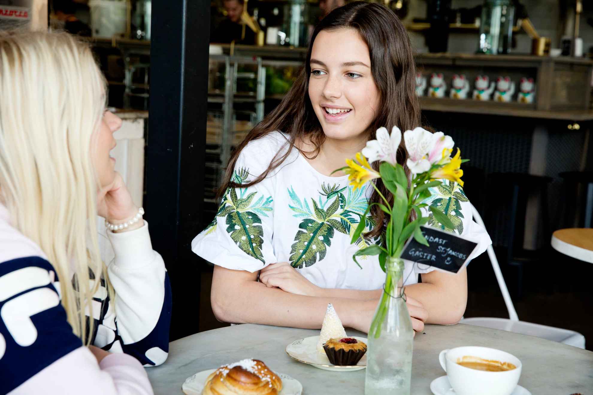 Zwei Frauen unterhalten sich und trinken dabei Kaffee und essen Fika Gebäck, wie eine Zimtschnecke und Muffins.