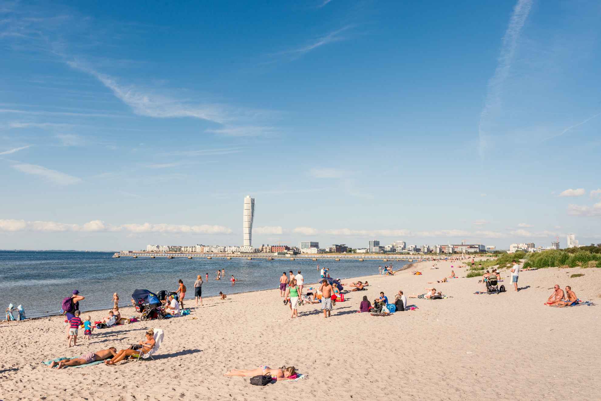 Menschen, die sich an einem Strand in Malmö vergnügen. Am Horizont ist das Gebäude "Turning Torso" zu sehen.