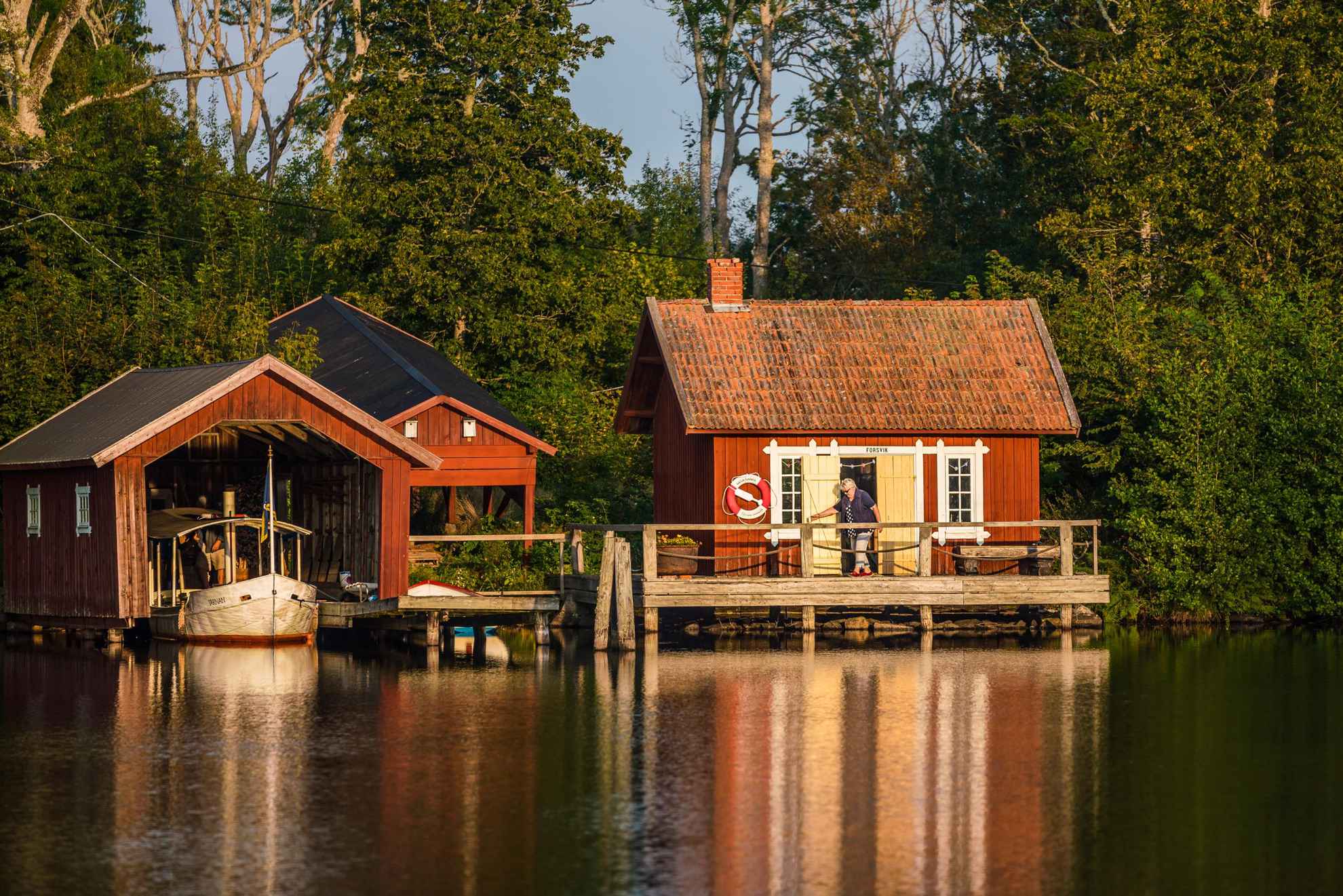 Eine Frau verlässt ein rotes Holzhaus, das am Wasser liegt. Neben dem Haus ist ein Bootshaus.