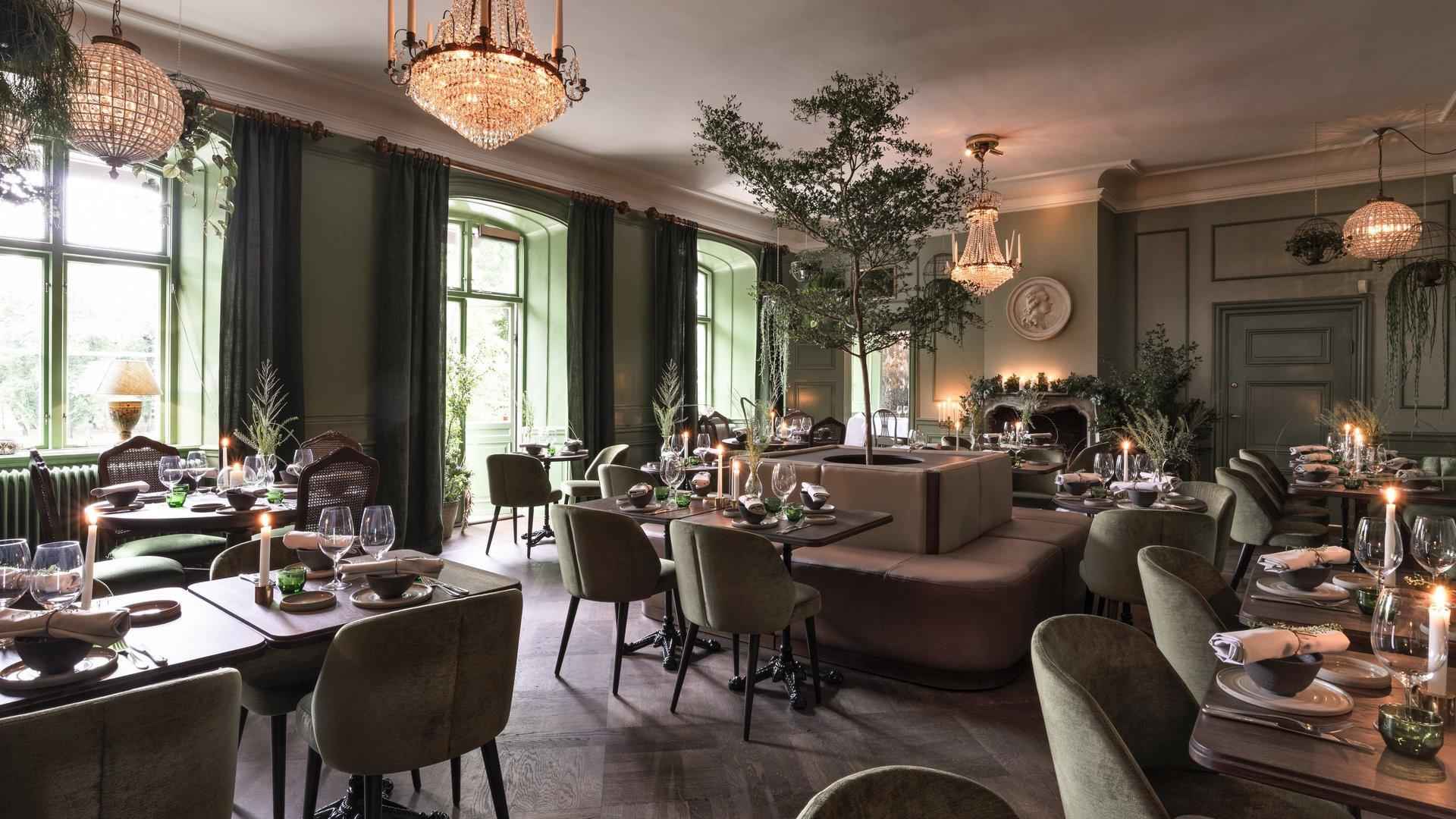 Ein großer Speisesaal, dekoriert mit Holztischen, grünen Stühlen und grünen Wänden. Von der Decke hängen Kronleuchter.