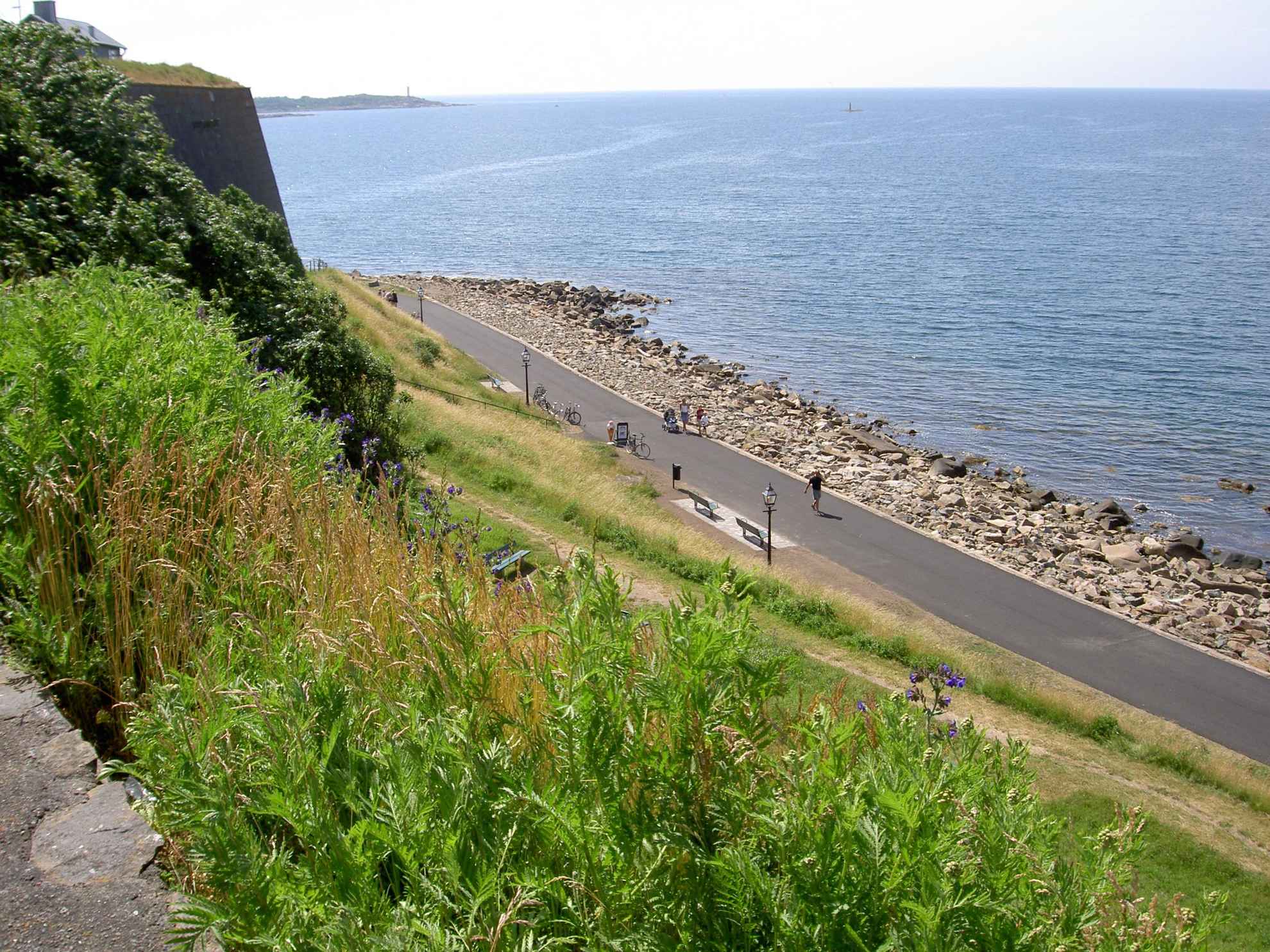 Eine Fahrradroute entlang der Uferpromenade, die vom Hügel oben aus gesehen wird. Menschen gehen auf der Straße und es gibt Fahrräder, die an der Seite geparkt sind. Grün im Vordergrund und das Meer im Hintergrund.