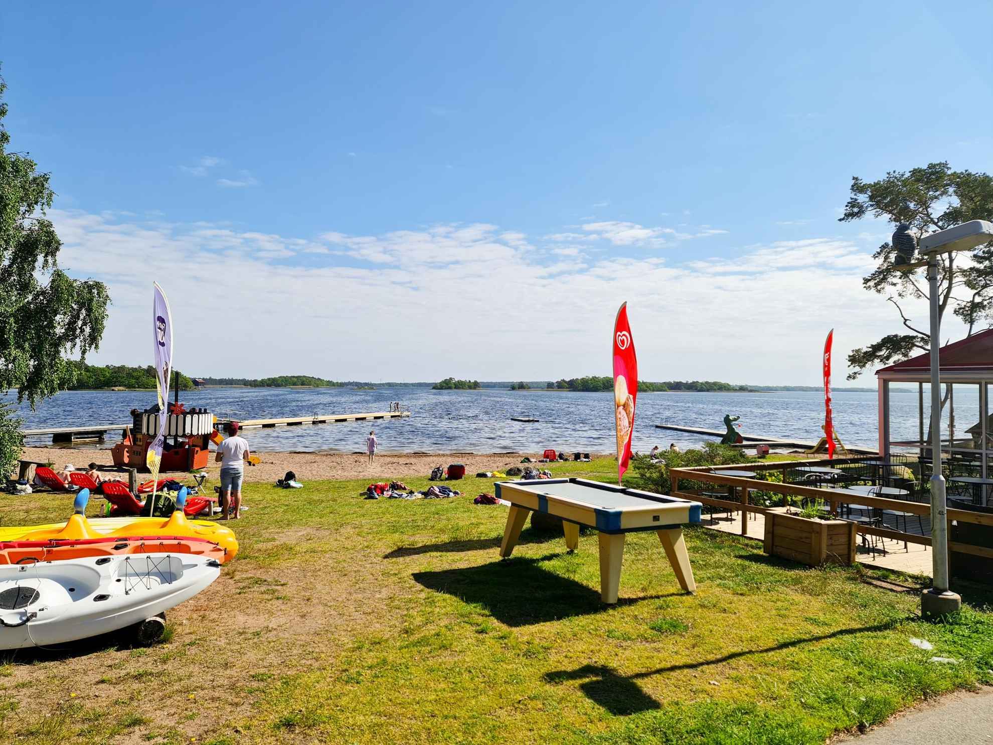 Ein paar Leute genießen einen sonnigen Tag an einem Strand mit zwei Stegen, Spielplätzen, Kanus und anderer Wassersportausrüstung. Auf der rechten Seite Teile eines Cafés.