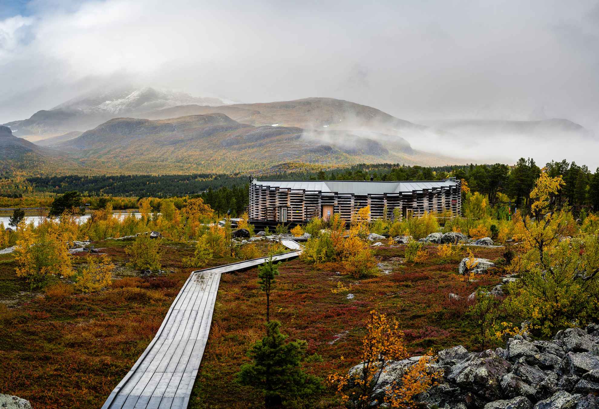Eine hölzerne Gehwegbrücke zum Gebäude des Naturum Laponia. Hinter dem Gebäude liegen Berge, die leicht von Nebel bedeckt sind.