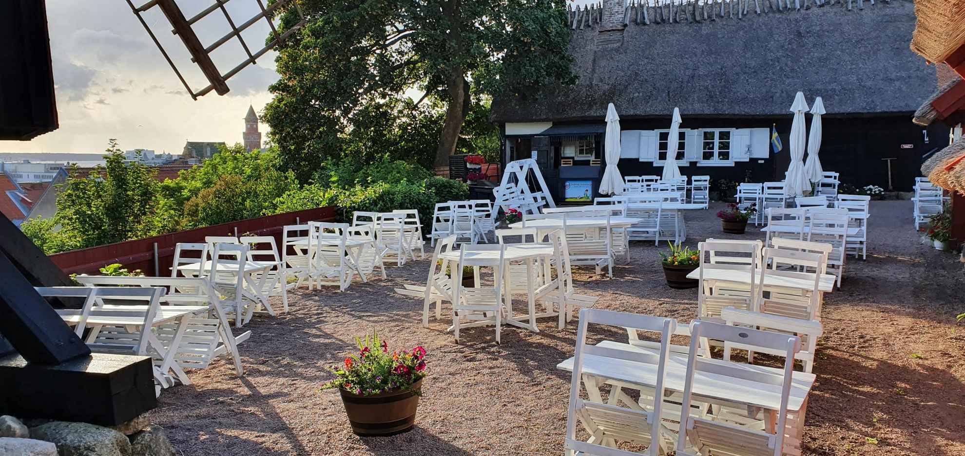 Ein Sitzbereich im Freien mit weißen Tischen und Stühlen in einem Restaurant und Café. Ein altes Haus und ein großer Baum im Hintergrund.