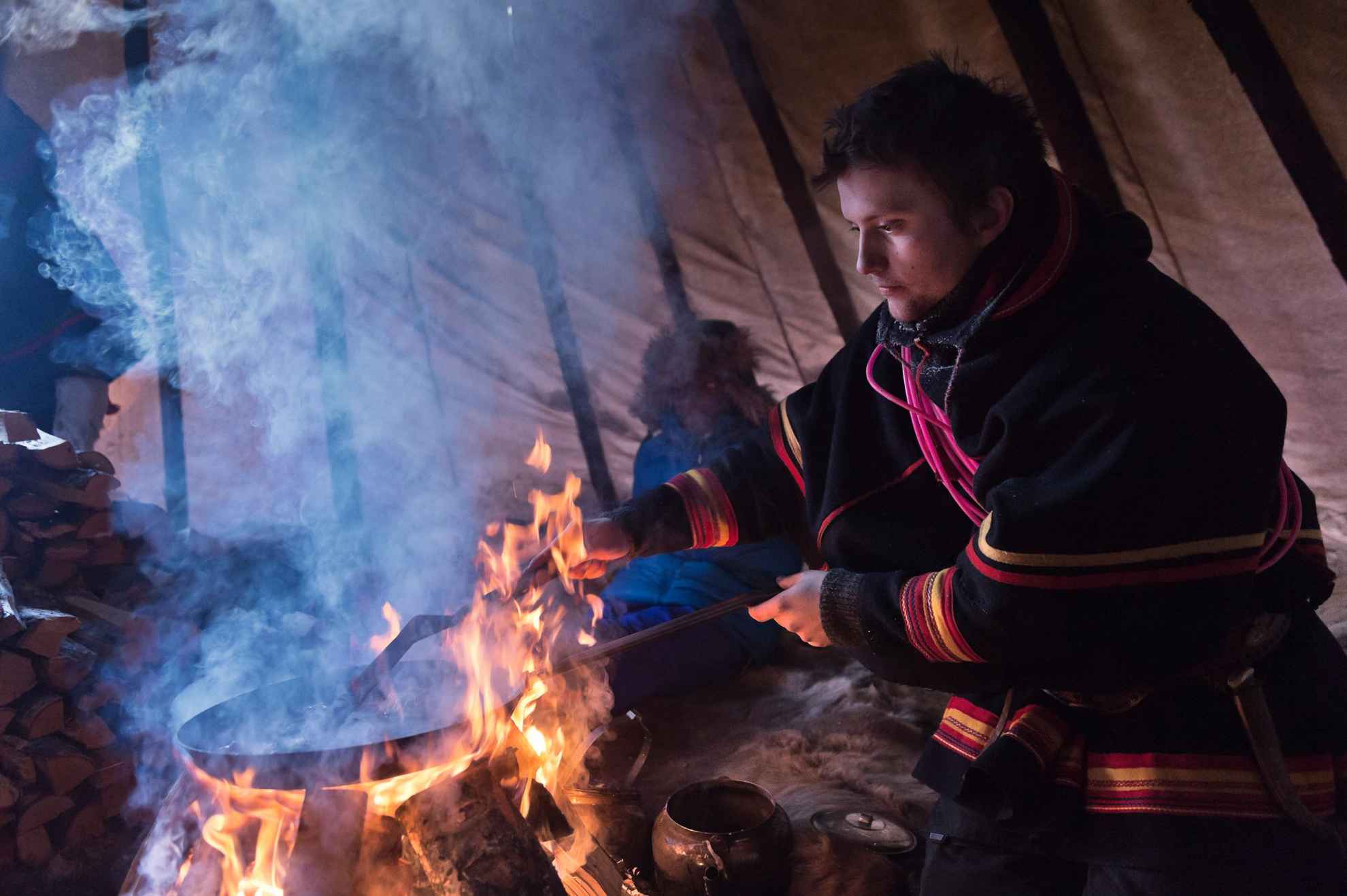 Ein Mann in samischer Tracht rührt in einer Pfanne, die über einem offenen Feuer hält. Alles in einem haushohen Zelt.