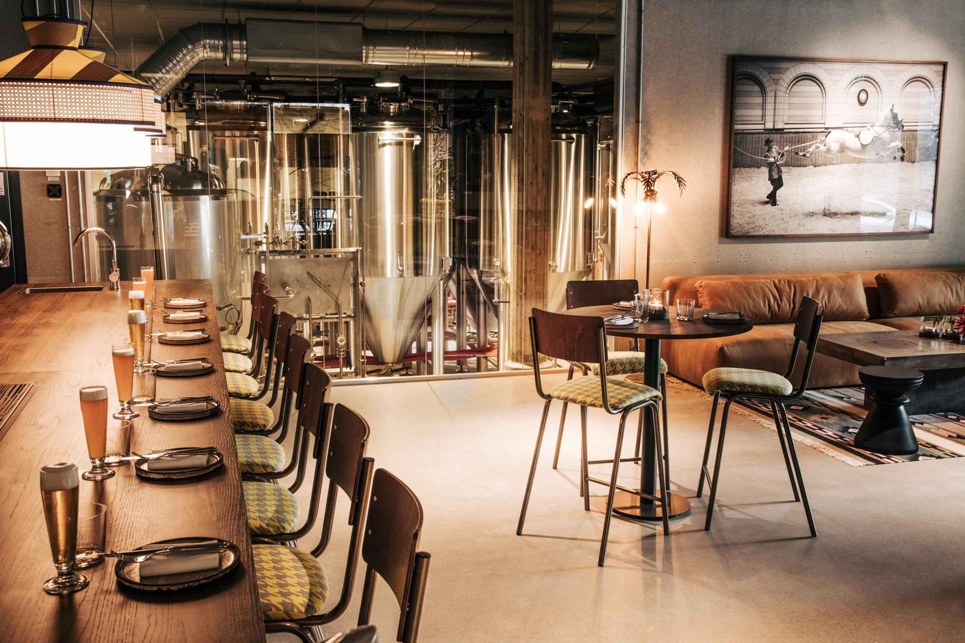 Im Inneren eines Restaurants ist die Theke auf der linken Seite mit Biergläsern bestückt, auf der rechten Seite steht ein gedeckter Tisch mit drei Stühlen und einem Sofa. Im Hintergrund kann man hinter einer Glaswand eine Brauerei sehen.