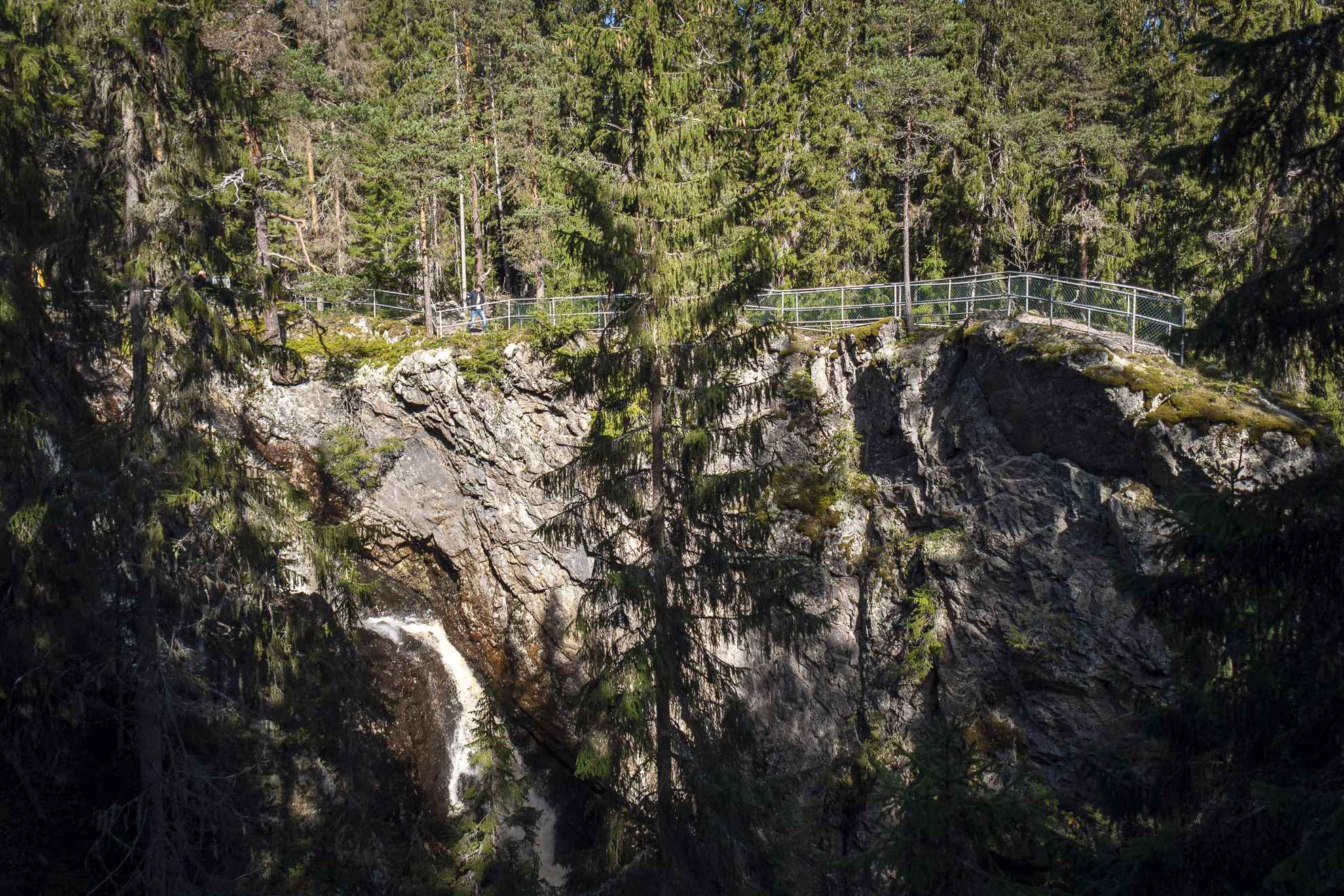 Ein Wasserfall umgeben von Wald. Ein Mann geht neben dem Zaun am Wasserfall.
